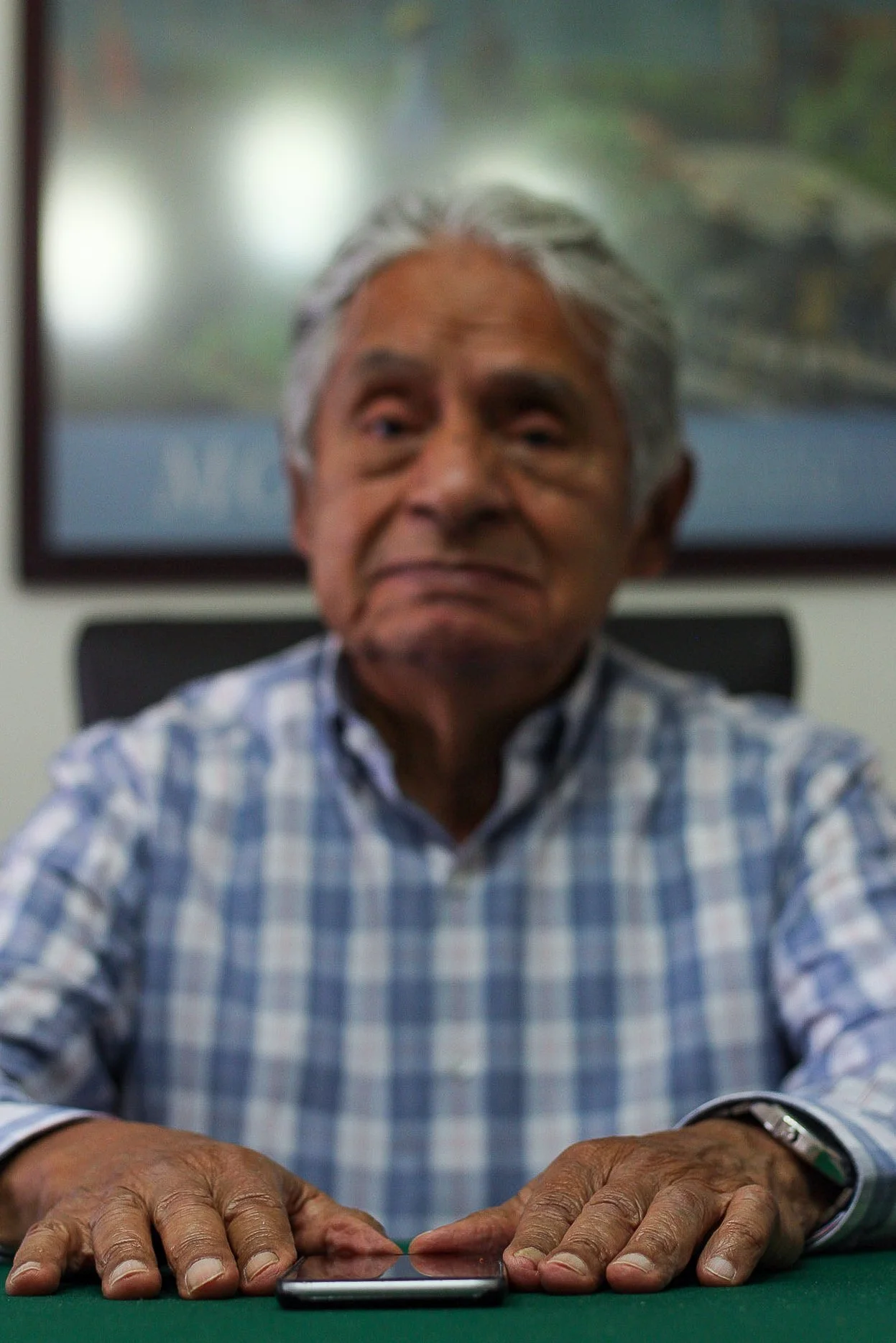 Hombre mayor con cabello gris, usando una camisa a cuadros, sentado en una mesa con un teléfono móvil frente a él.
