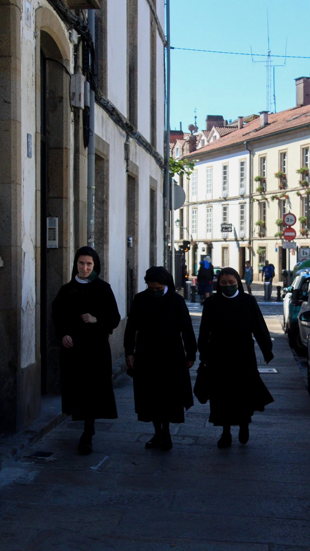 Tres monjas caminando juntas en una calle urbana, con edificios antiguos y un cielo despejado al fondo. Todas llevan hábito negro y mascarillas negras.