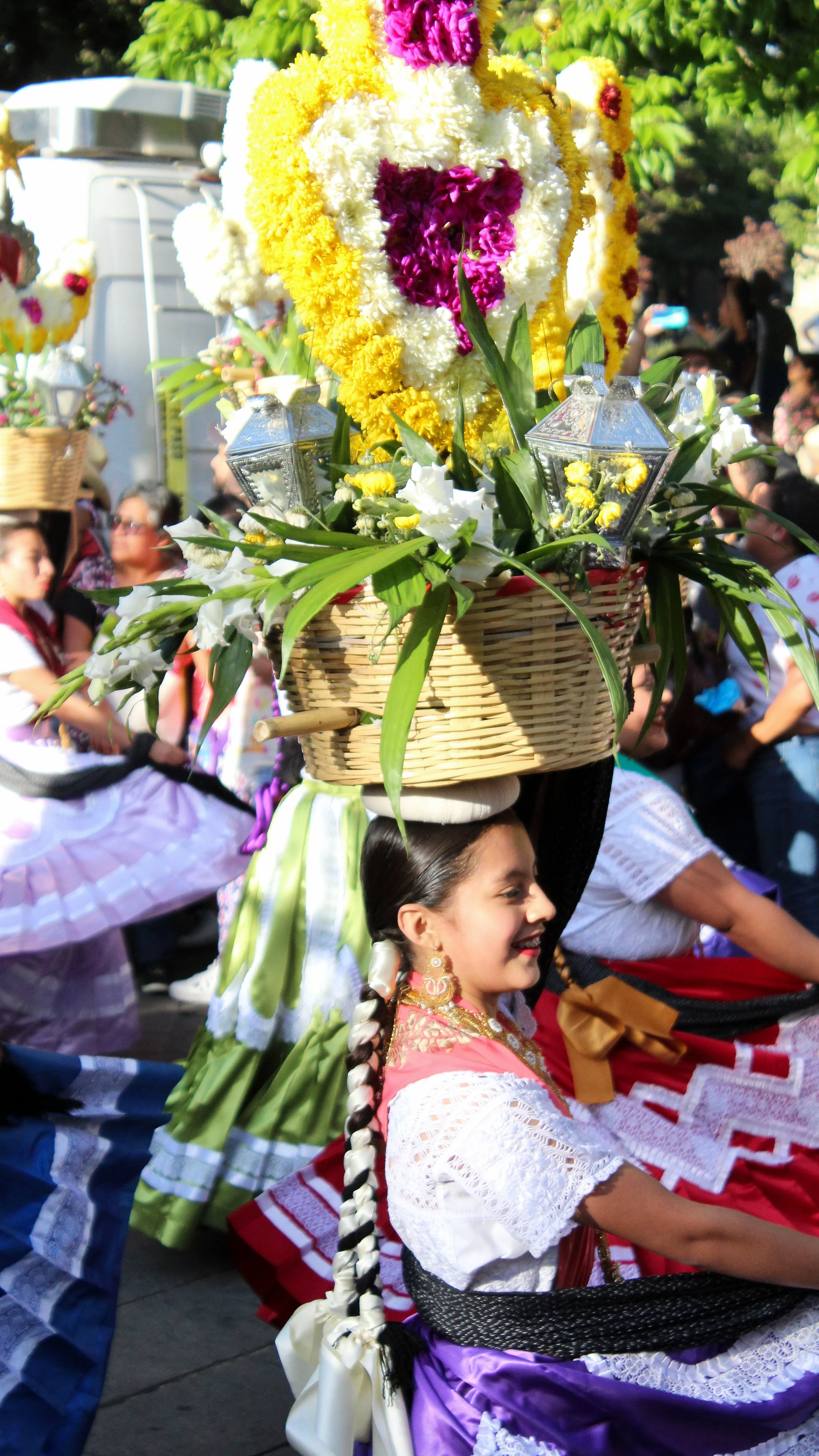 Mujer vestida con ropa tradicional, sonriendo, lleva en la cabeza una canasta con flores y velas, participan en una celebración o desfile.