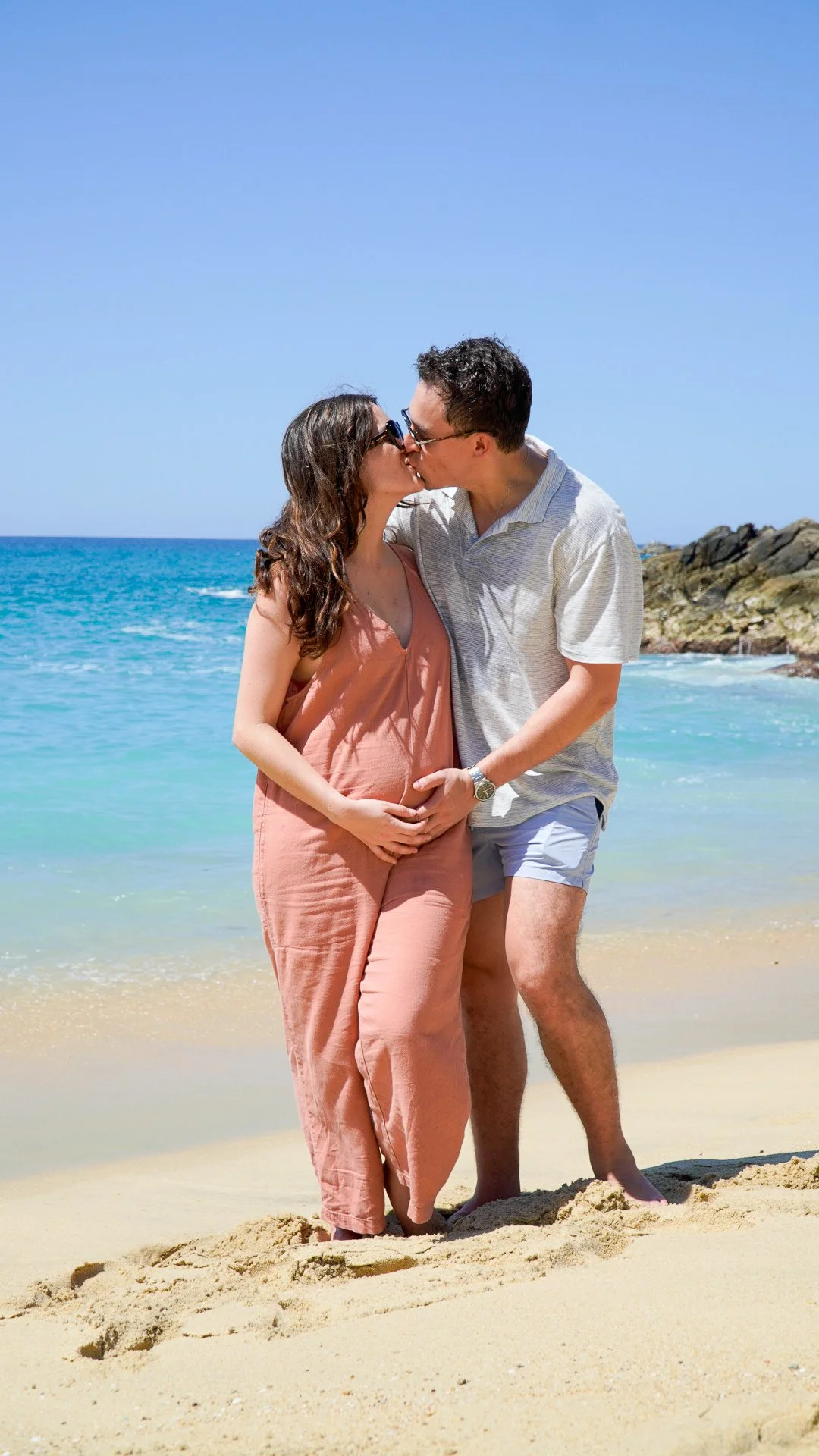 Una pareja se besa en la playa bajo un cielo azul con agua cristalina y rocas al fondo.