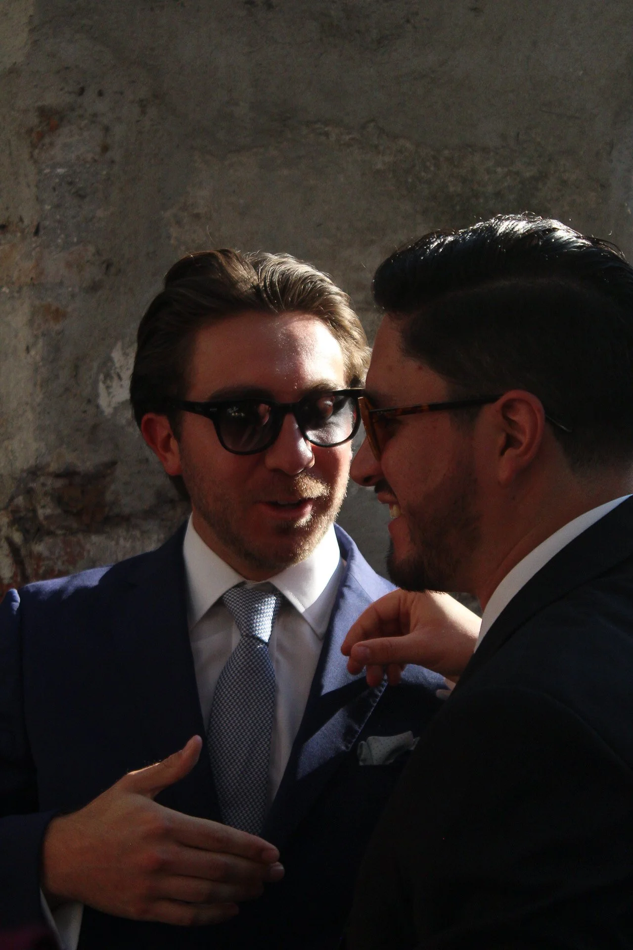 Dos hombres con trajes oscuros y gafas de sol en una conversación cercana, en un fondo de muro de ladrillo en tonos grises.
