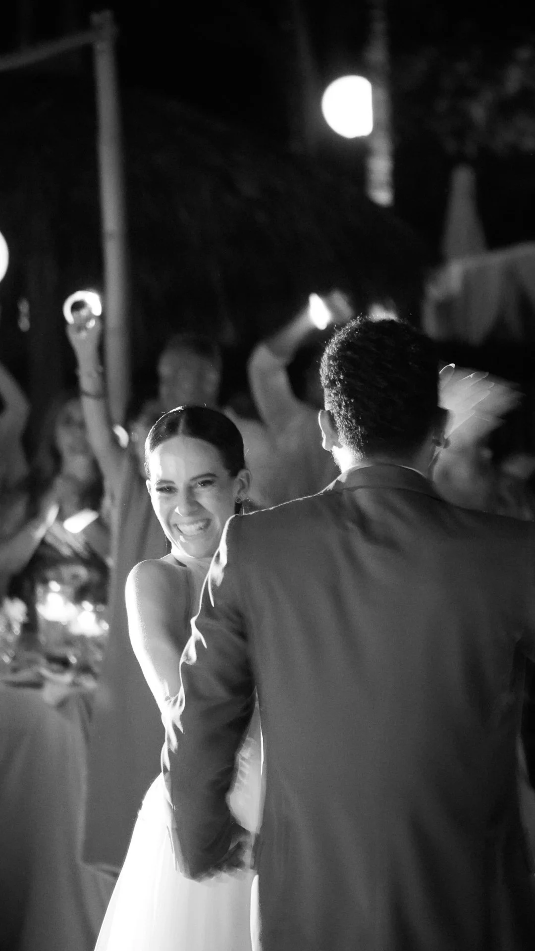 Una pareja bailando en una boda. La novia sonríe con alegría mientras mira al novio, en un entorno con personas y luces en el fondo.