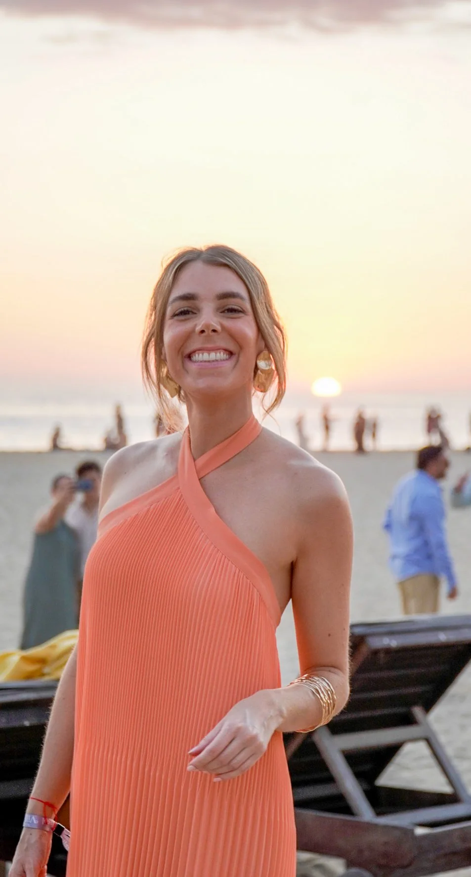 Mujer sonriendo en la playa durante el atardecer, vestida con un vestido naranja, con personas al fondo y el mar.