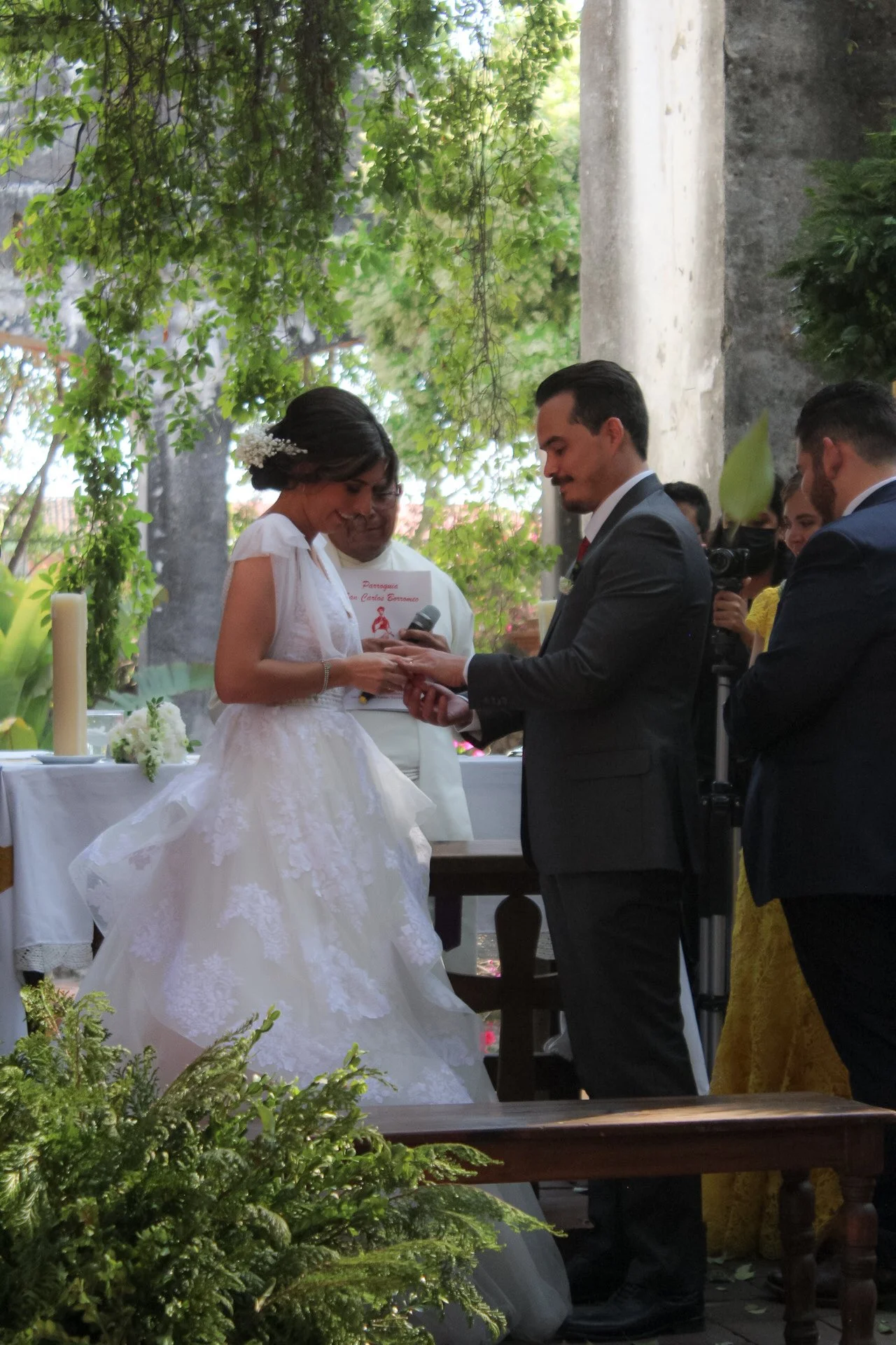 Pareja casándose en una ceremonia al aire libre con plantas verdes y árboles, con la novia en vestido blanco y el novio en saco gris.