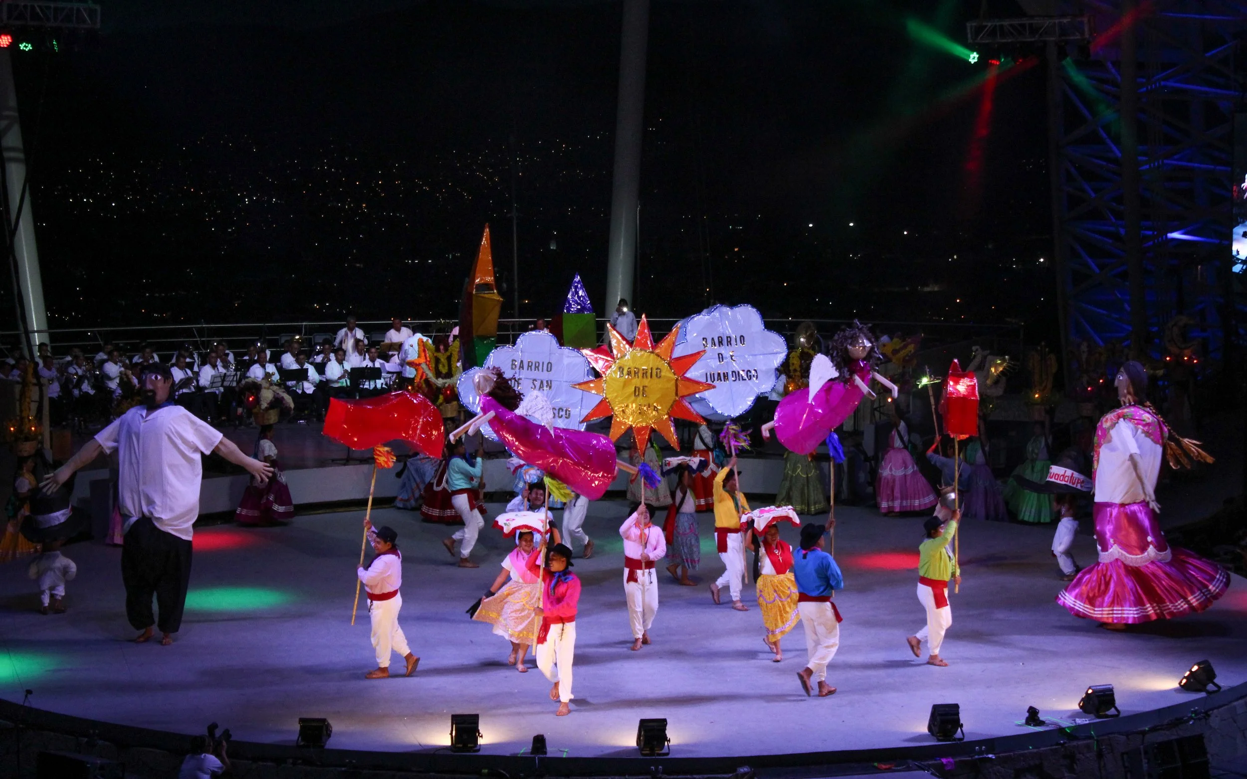 Grupo de personas bailando en un escenario con vestuarios tradicionales, llevando globos y sombreros decorativos, con una orquesta tocando en el fondo y luces de colores en la noche.