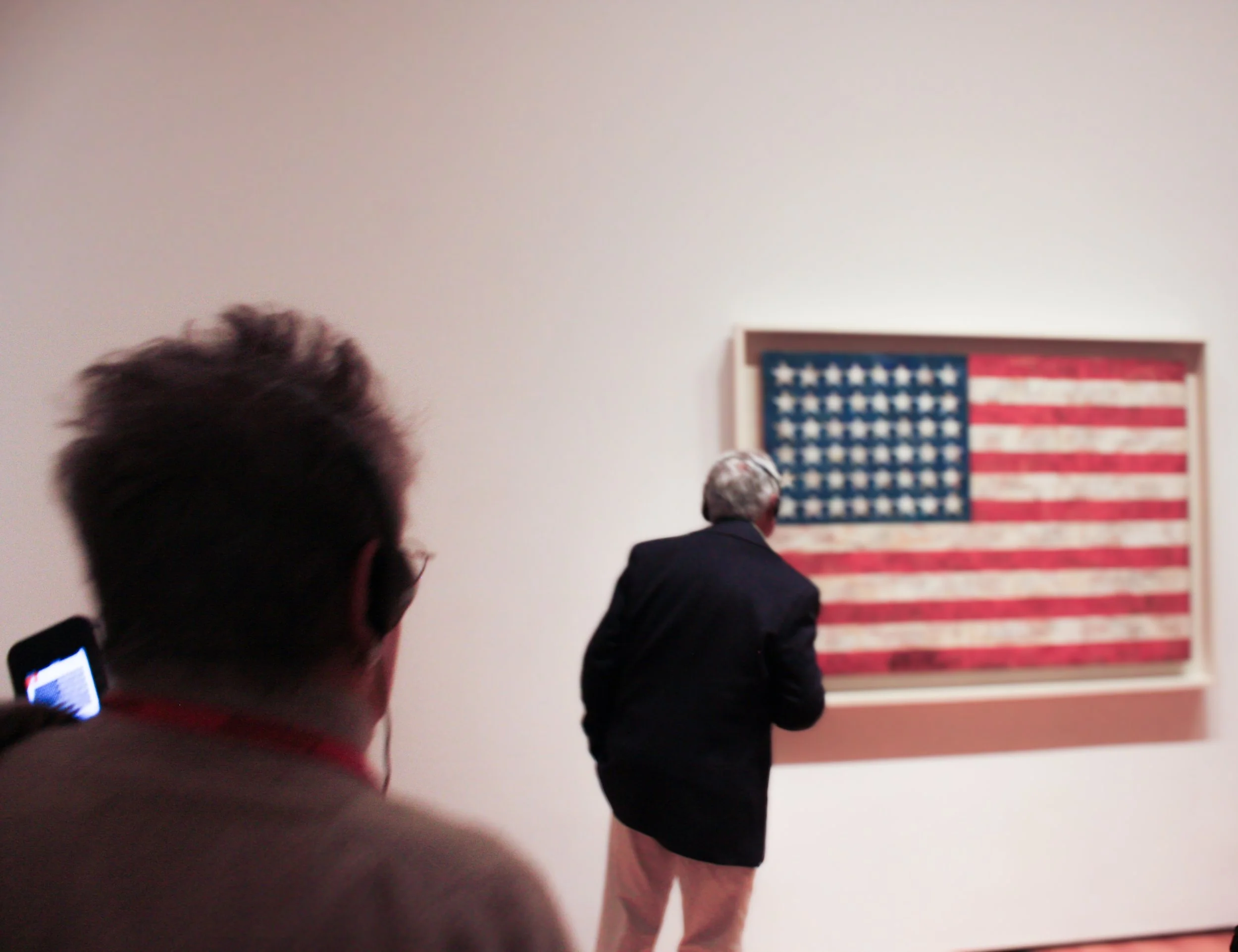 Dos hombres observan una obra de arte en una galería, con una bandera de Estados Unidos decorativa en la pared.