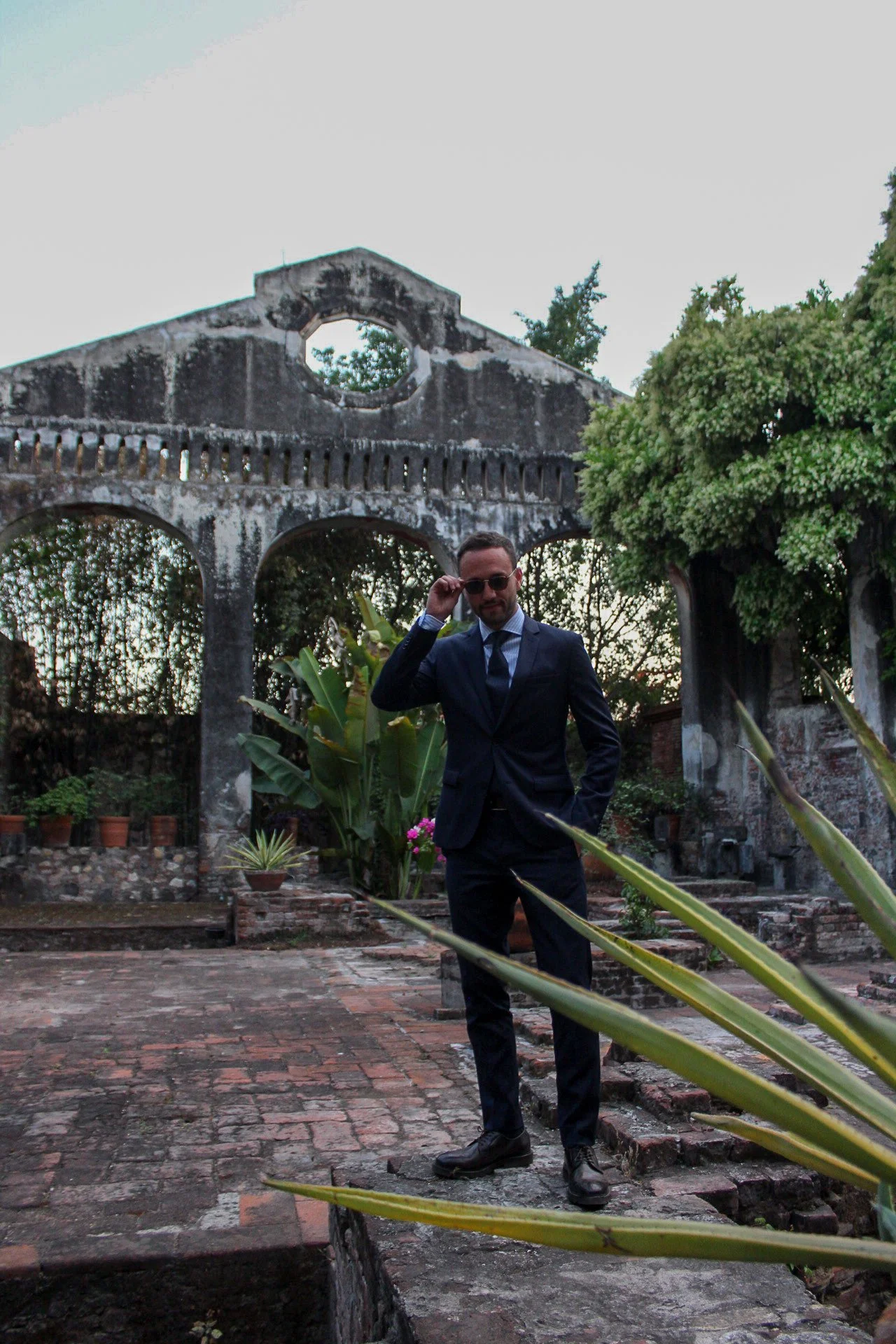 Hombre vestido con traje oscuro, gafas de sol y corbata, en un jardín con plantas verdes y una estructura antigua de piedra en el fondo, al atardecer.