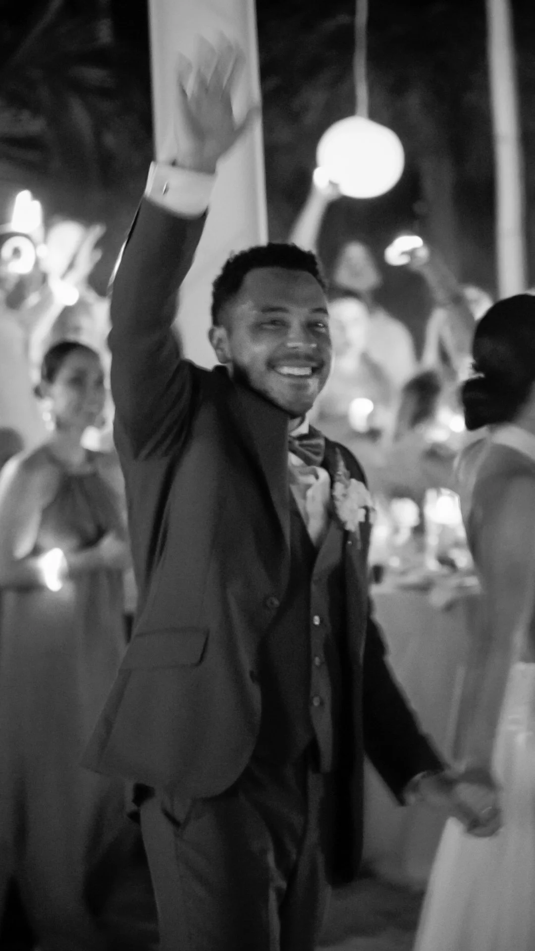 Un hombre sonriendo, vestido con traje formal y una pajarita, en un evento social, saludando con la mano en señal de saludo o despedida, en un ambiente con muchas personas y decoraciones luminosas.