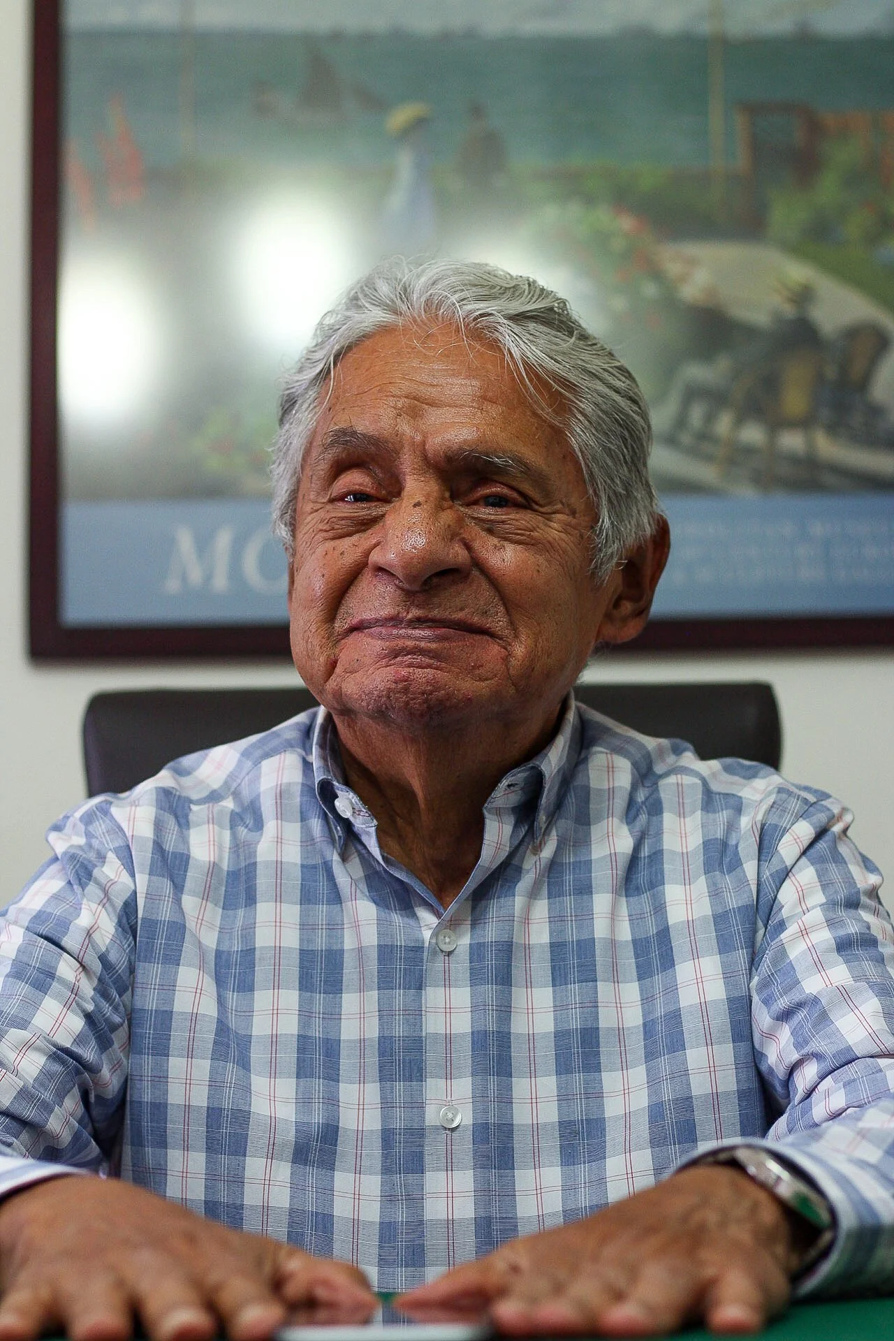 Un hombre mayor con cabello gris y expresión serena, usando una camisa a cuadros, sentado en una oficina con un cuadro de fondo.