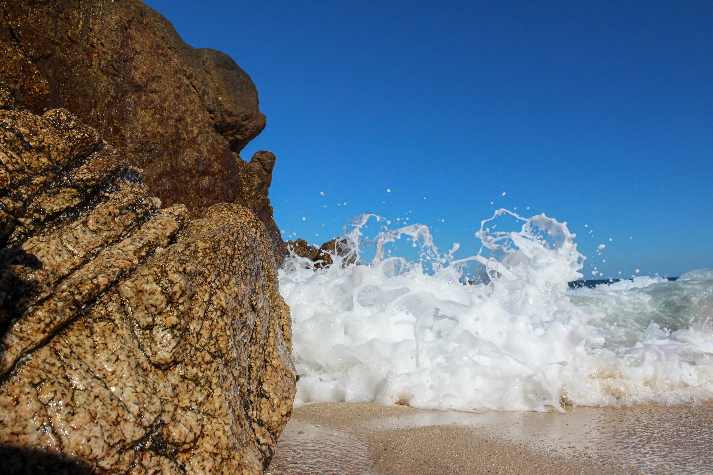 Olas rompen contra rocas en la orilla de una playa, fondo de cielo despejado, día soleado.