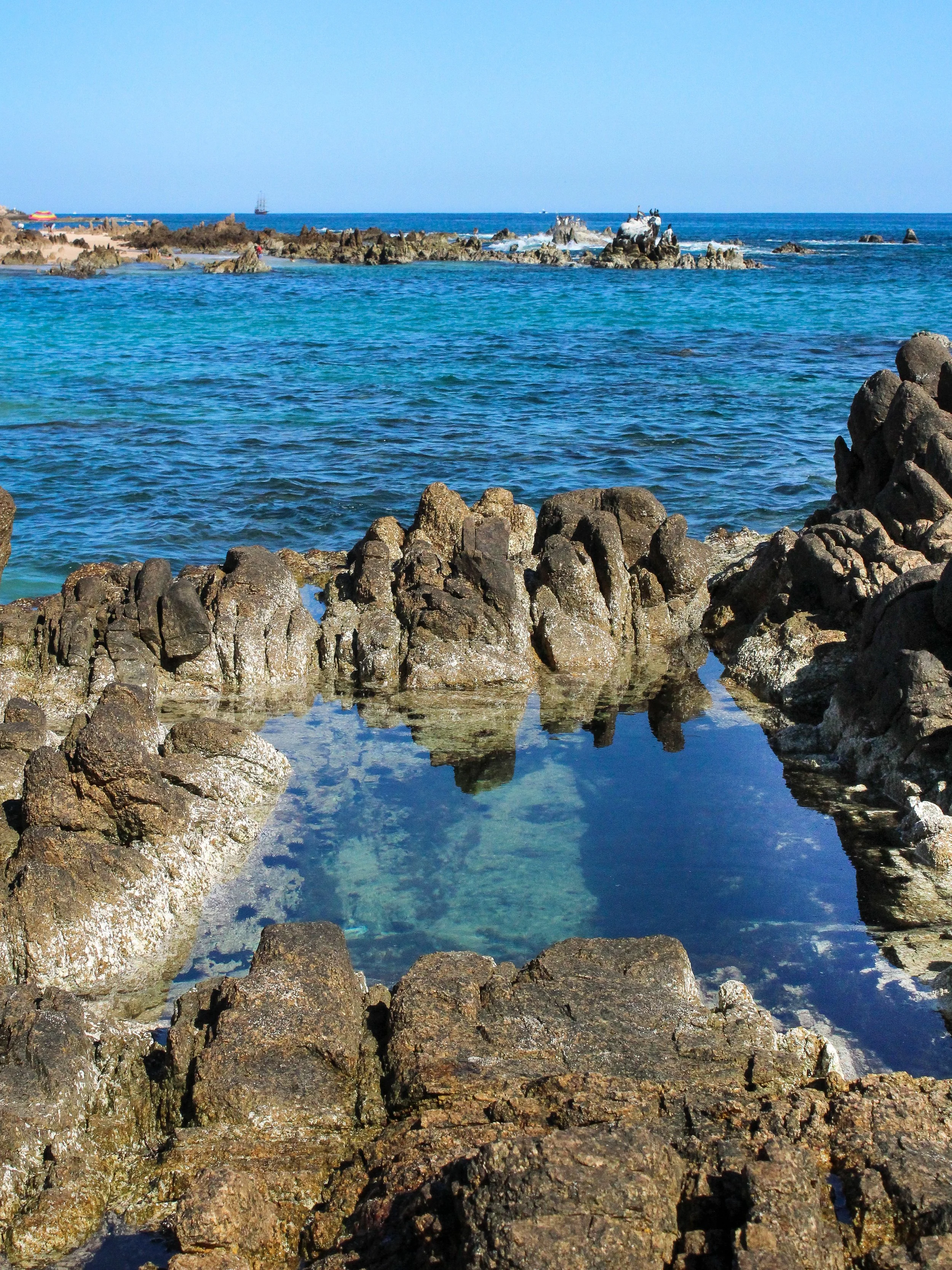 Costa de mar con rocas y un estanque natural de agua en primer plano, y mar azul con formaciones rocosas en el fondo bajo cielo despejado.