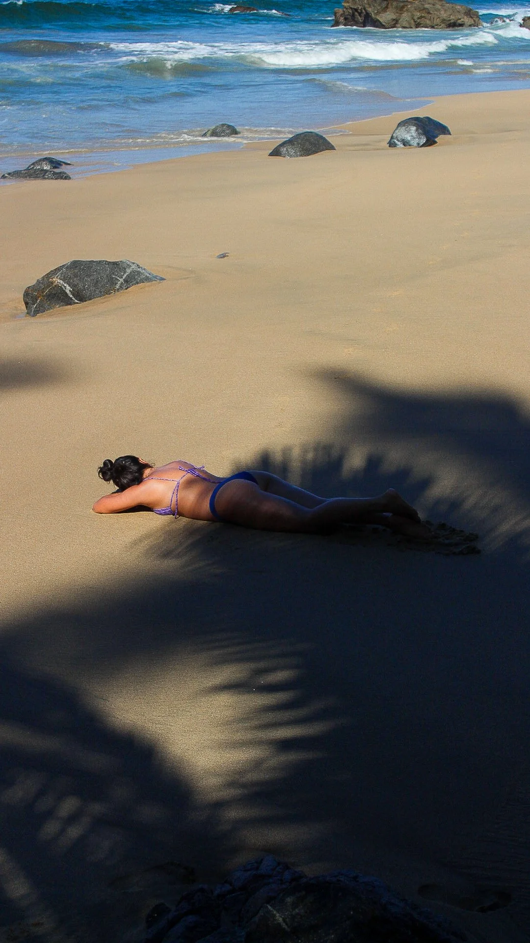 Mujer descansando en la playa acostada sobre la arena, sombra de palmera en primer plano, en fondo olas del mar y rocas en la orilla.