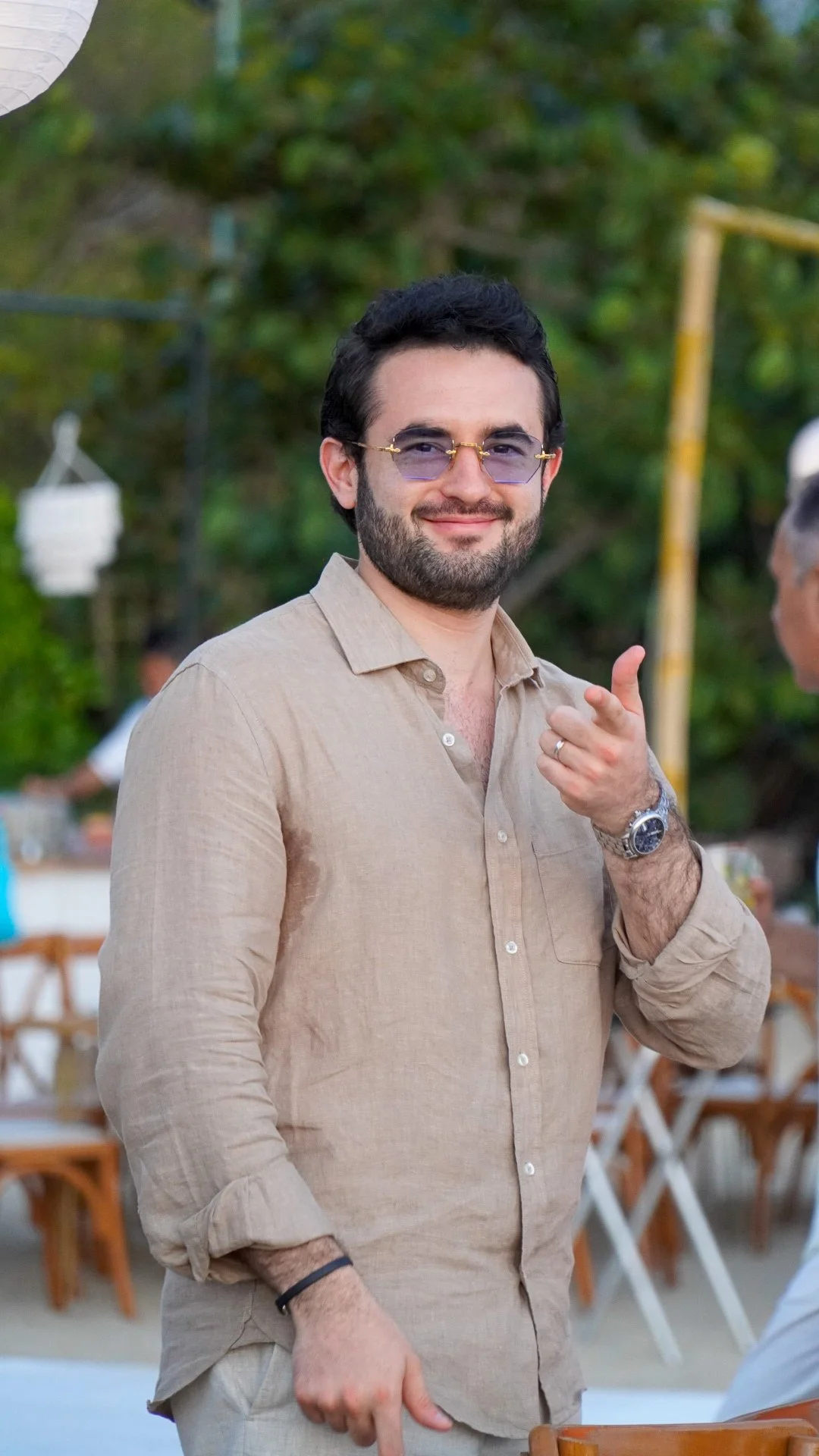 Hombre con barba, gafas de sol, camisa beige, sonriendo y señalando hacia la cámara en fondo exterior con árboles y sillas de madera.