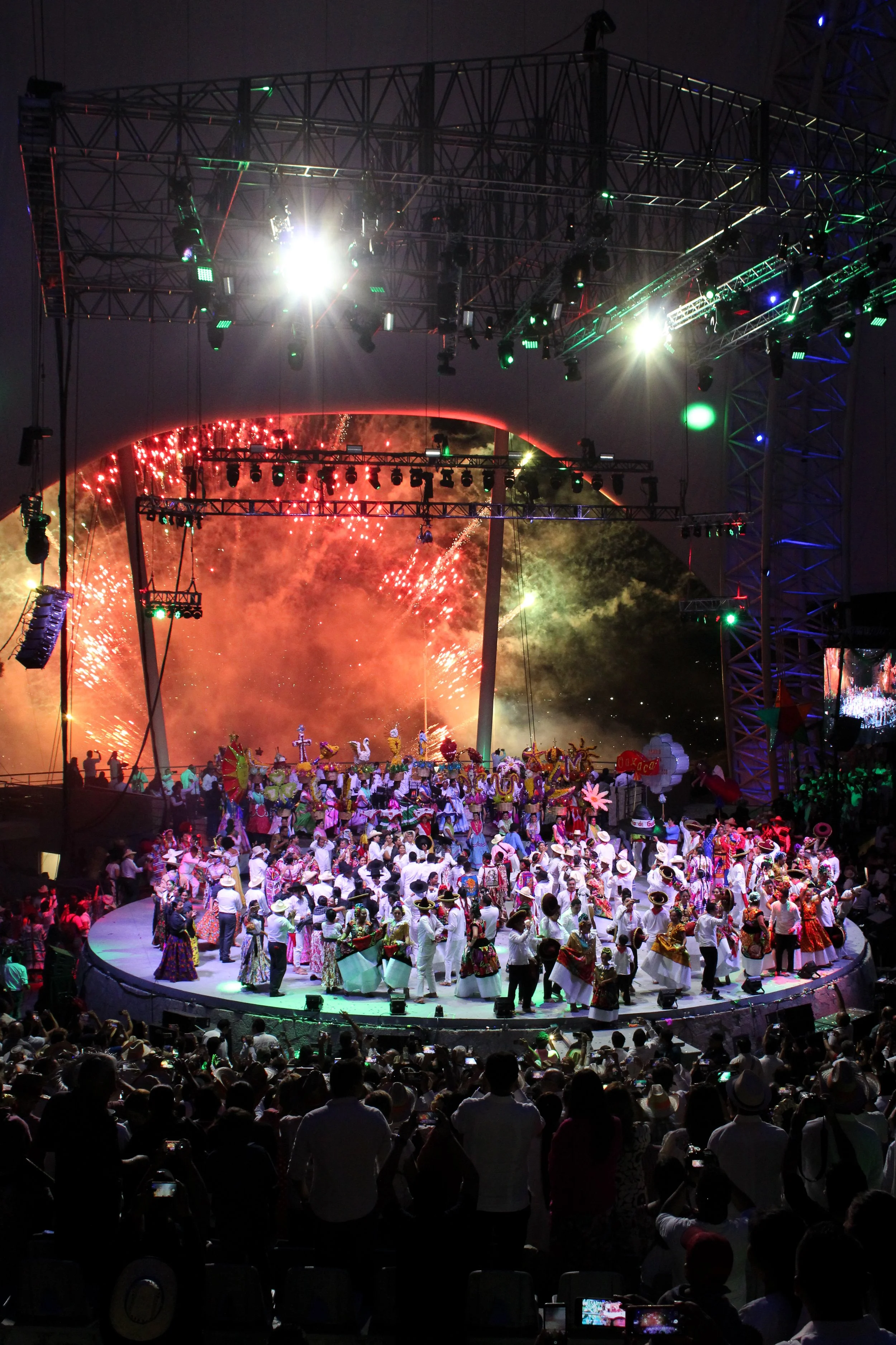 Espectáculo cultural con personas vestidas con trajes tradicionales en un escenario, con fuegos artificiales en el fondo y un público observando.