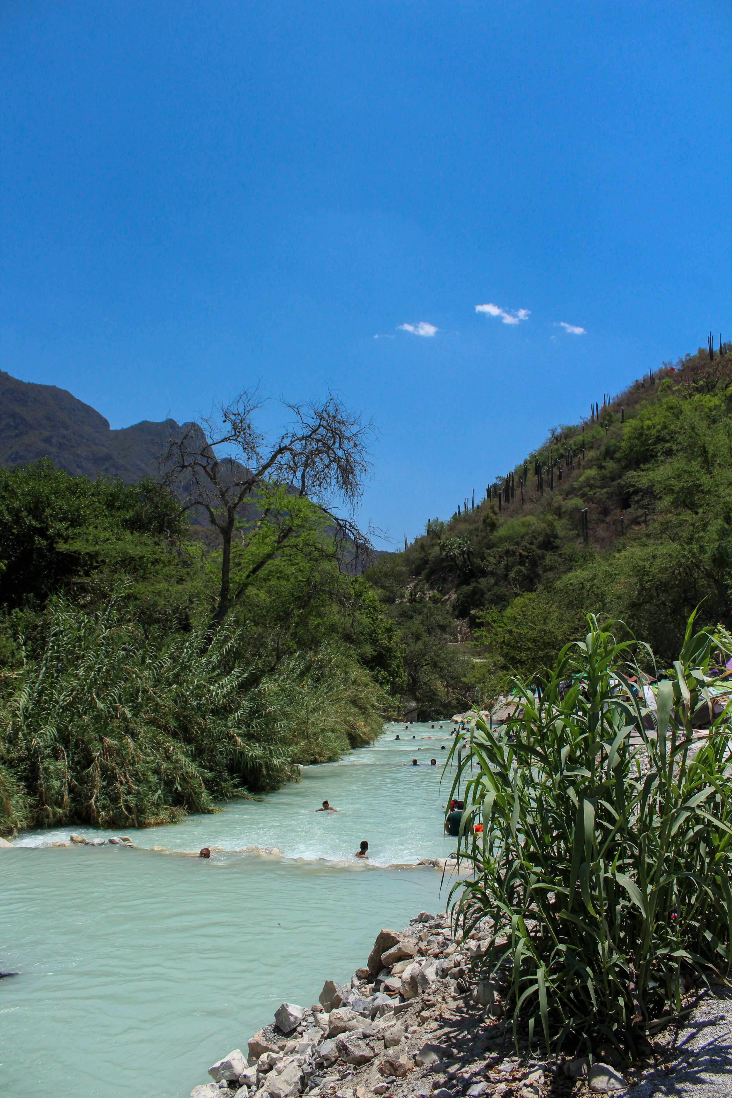 Vista de un río turquesa en un cañón rodeado de vegetación y montañas, con varias personas nadando y un cielo azul claro.