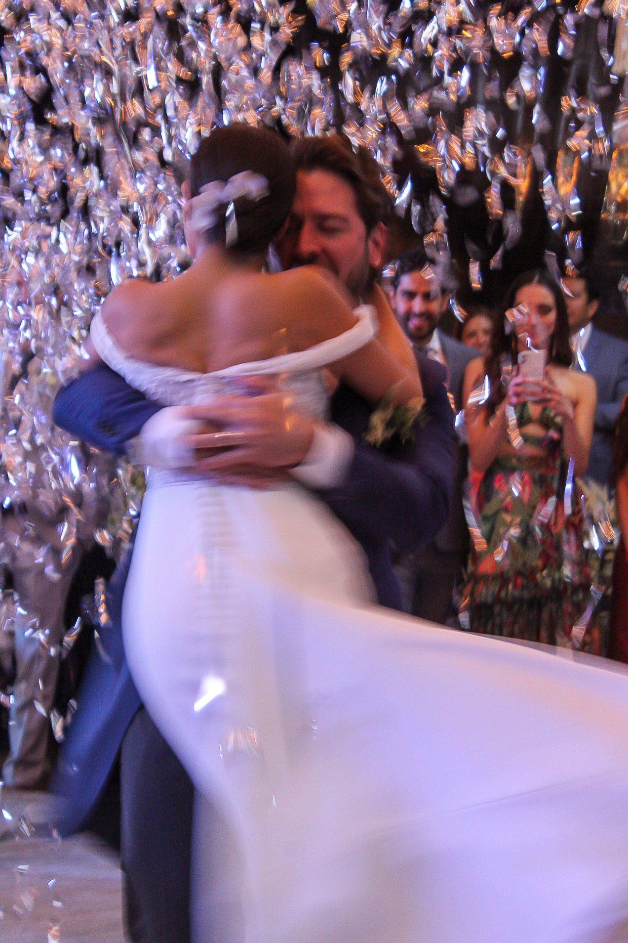 Pareja bailando en una boda con confeti en el fondo, mientras varias personas los observan y toman fotos.