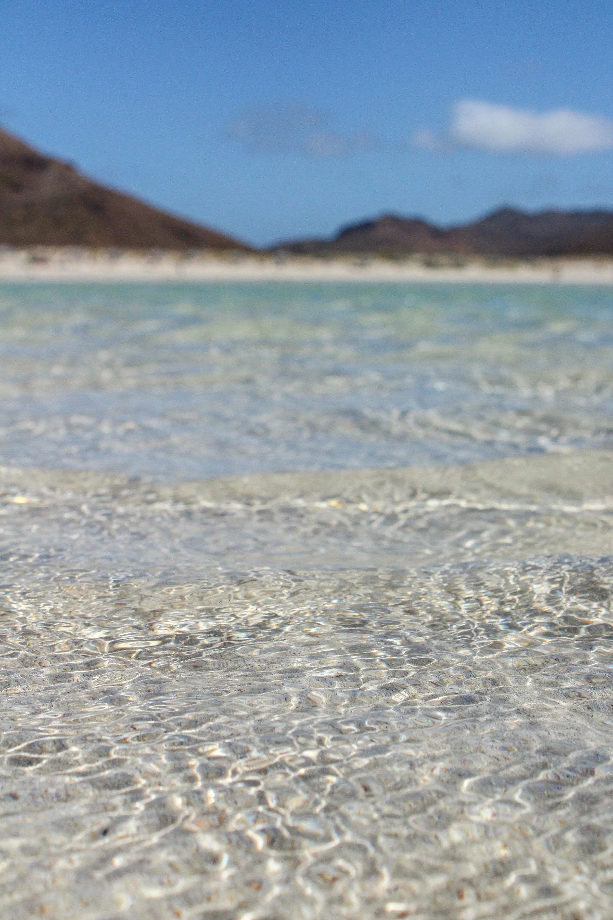 Vista del mar con agua cristalina en la orilla, montañas a lo lejos y cielo azul con algunas nubes.
