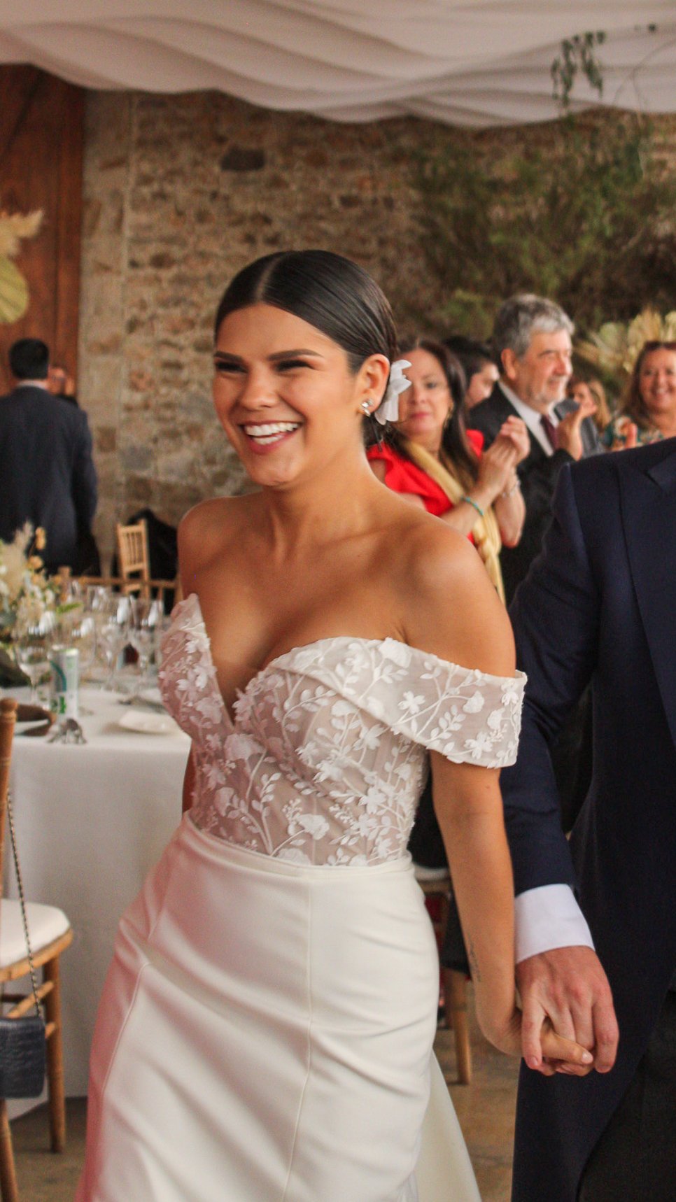 Mujer en vestido de novia sonriendo en una boda, rodeada de invitados en un salón decorado con flores y paredes de piedra.