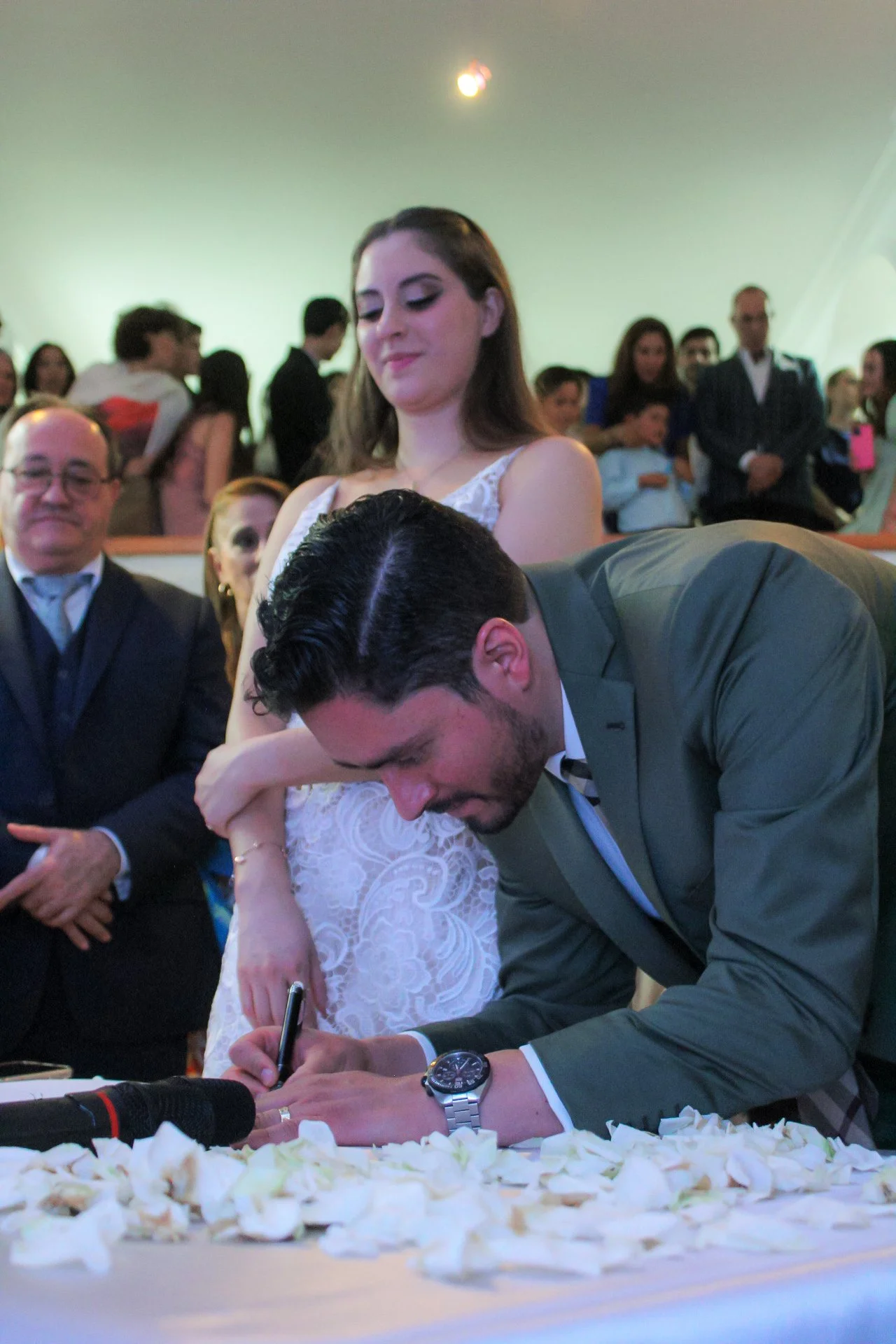 Hombre firmando un documento en una ceremonia de boda, con mujer en vestido blanco de pie detrás y varias personas observando en el fondo.