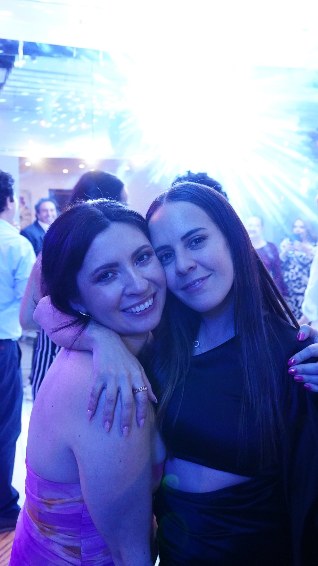 Dos mujeres sonriendo y abrazándose en una fiesta con luces de discoteca en el fondo.