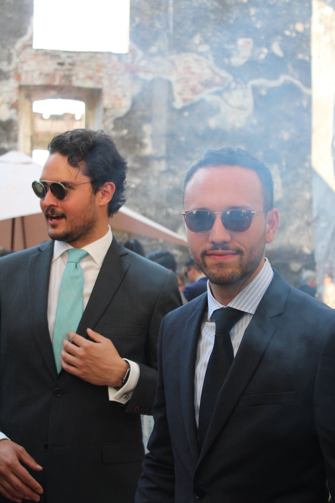 Dos hombres con trajes oscuros y gafas de sol en un evento social, con fondo de muro de piedra y personas en el lugar.