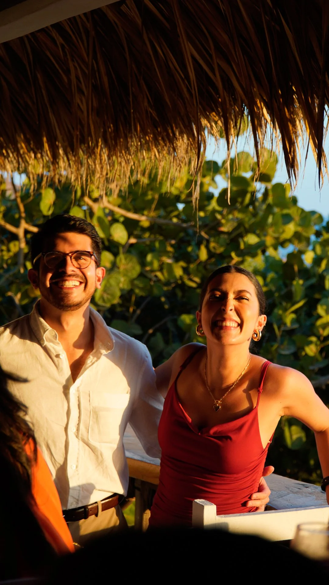 Dos personas sonrientes, un hombre con gafas y camisa color crema, y una mujer con vestido rojo y joyas, en un entorno tropical con vegetación baja y un techo de palma. El atardecer ilumina la escena.