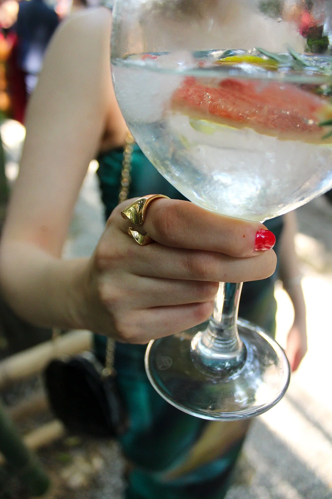 Mano sosteniendo una copa de cóctel con hielo, rodajas de cítricos y hierbas aromáticas, en un entorno al aire libre, con algunas personas de fondo.