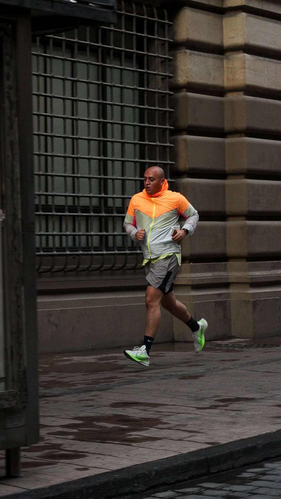 Persona corriendo en la calle, viste ropa deportiva de colores naranja, gris y verde, lleva un teléfono en la mano y usa zapatillas blancas y negras.