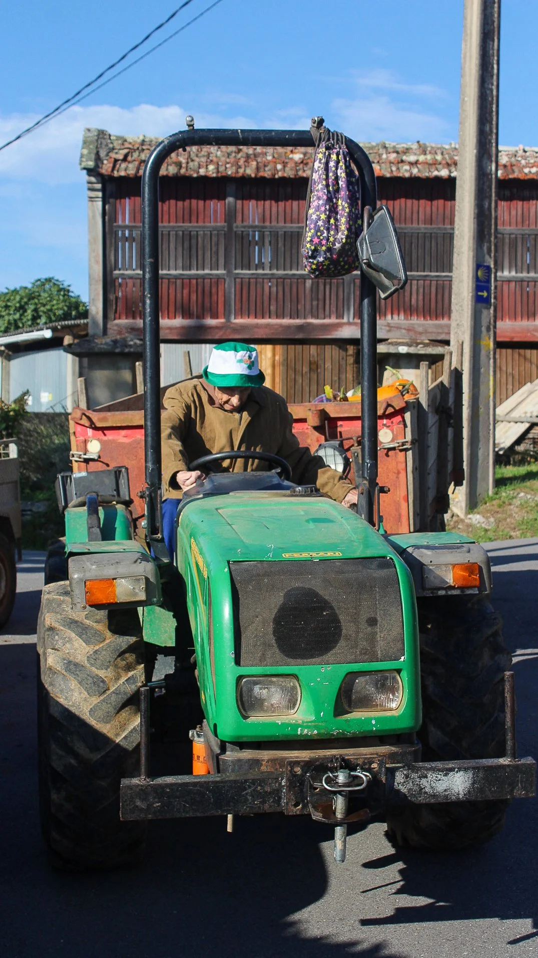 Un anciano sentado en un tractor verde en una calle, con una bolsa colgada en la barra y un saco en el respaldo, en un entorno rural con casas y un cielo azul.