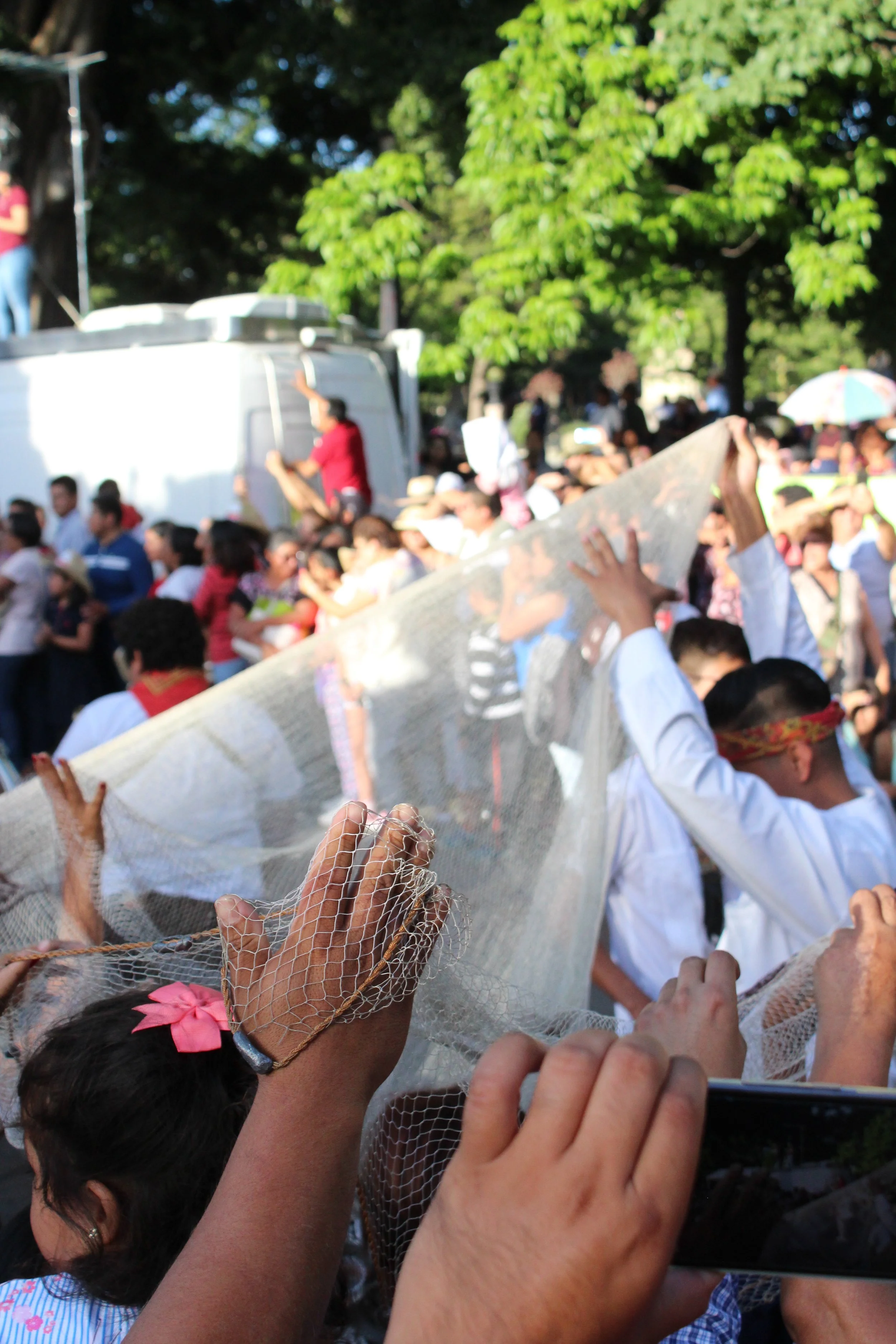 Personas participando en una celebración tradicional con una red de malla, en medio de un gran grupo de gente durante el día, en un entorno al aire libre con árboles verdes.