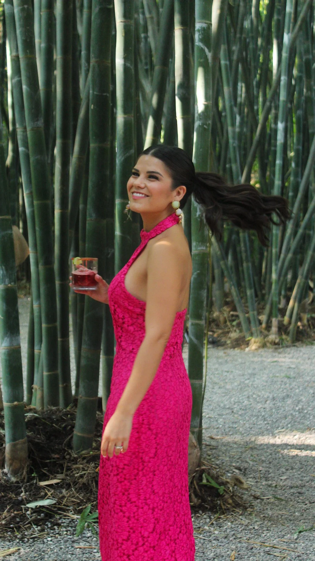 Mujer sonriendo, vestida con vestido rosa, sosteniendo una copa, en un entorno de bambú.