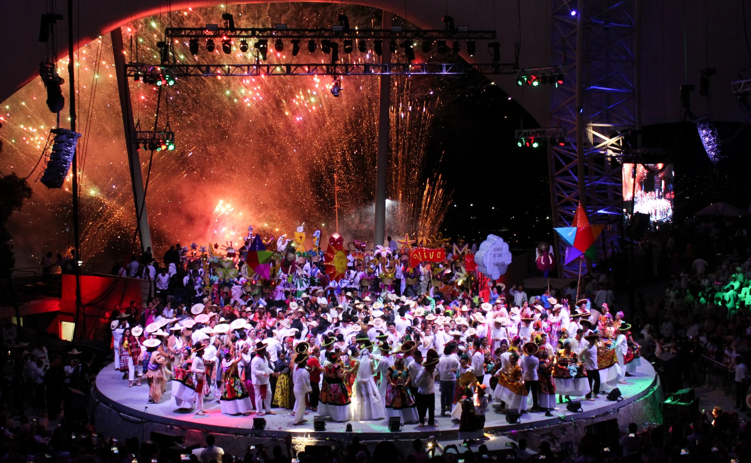 Celebración tradicional con personas vestidas con trajes folclóricos en un escenario, fuegos artificiales en el fondo, y decoraciones coloridas, en un ambiente festivo nocturno.