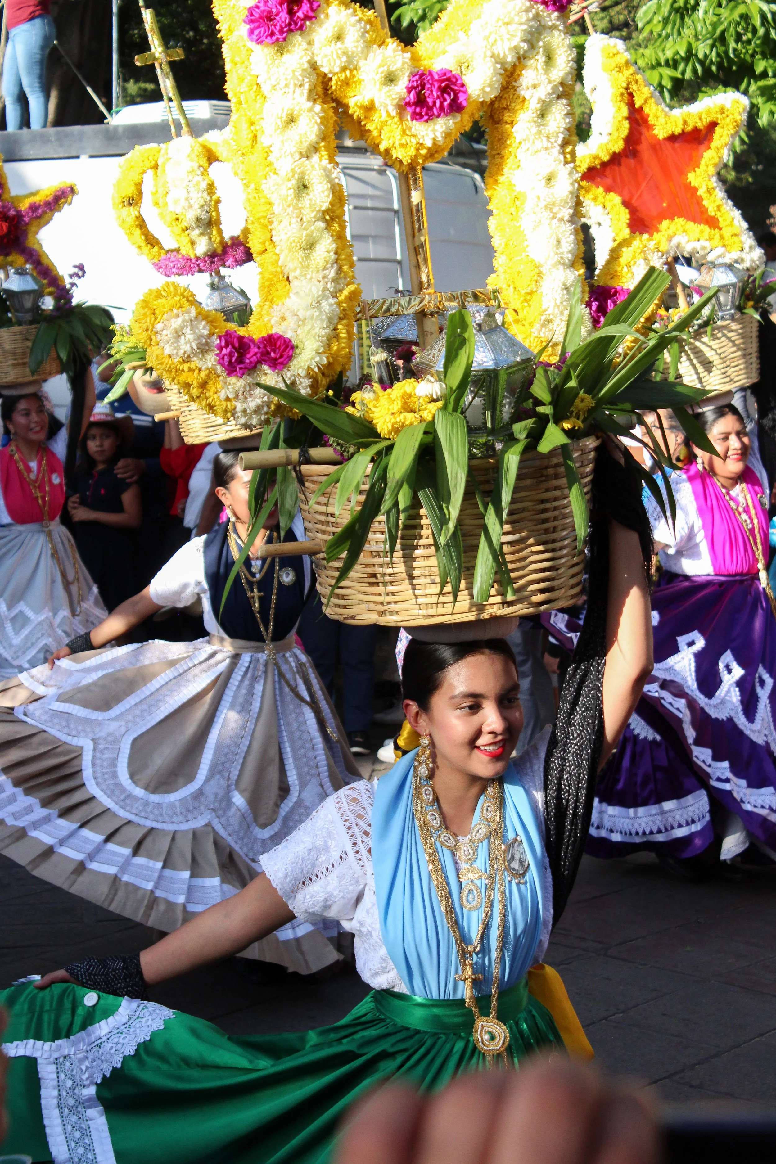 Mujer vestida con ropa tradicional mexicana participando en un desfile, llevando en la cabeza una canasta llena de flores coloridas, en un entorno festivo con otras personas también vestidas con trajes típicos.