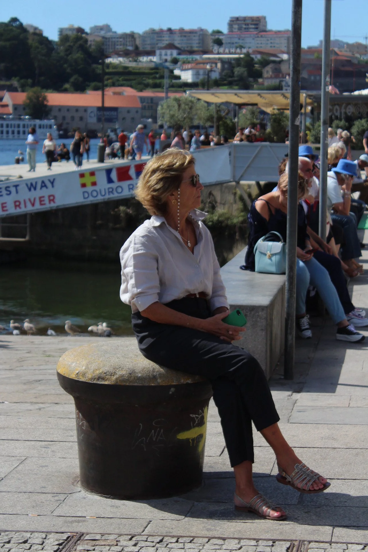 Mujer sentada en un banco junto a un río con varias personas en el fondo y edificios en la colina.