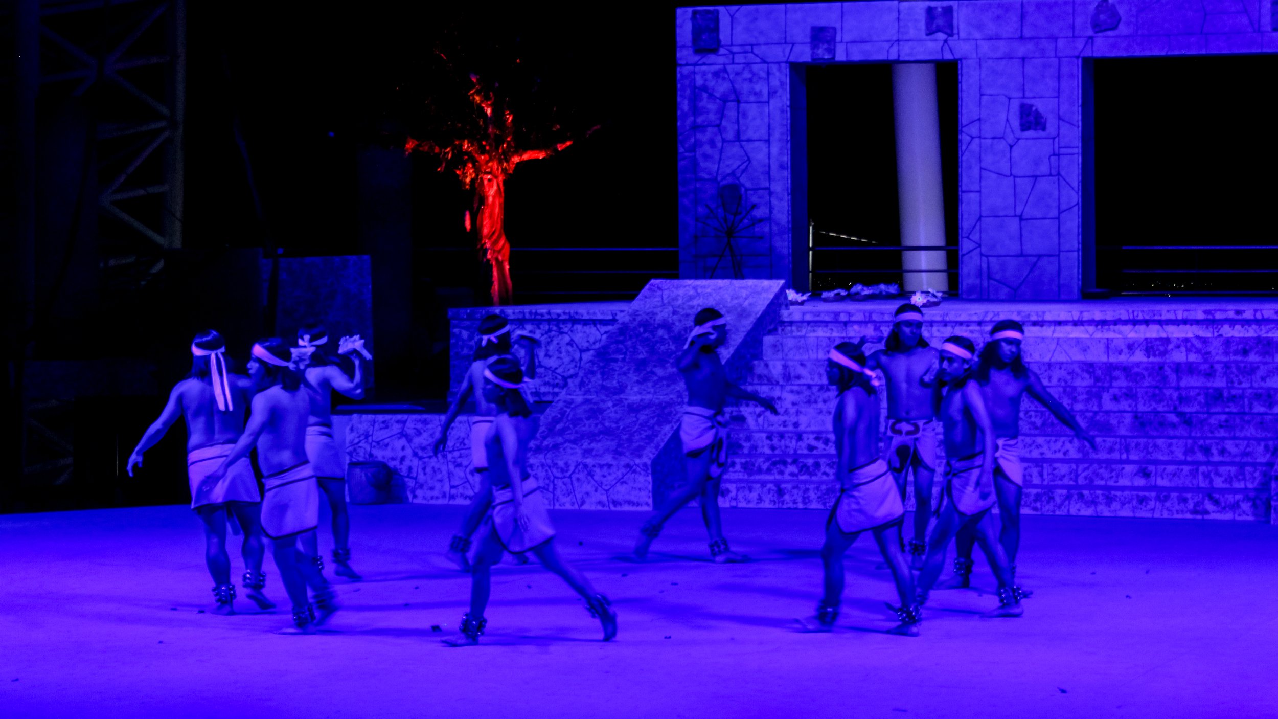 Grupo de niños realizando una danza en un escenario con iluminación azul, vestimenta tradicional y accesorios en la cabeza, en un show cultural al aire libre por la noche.