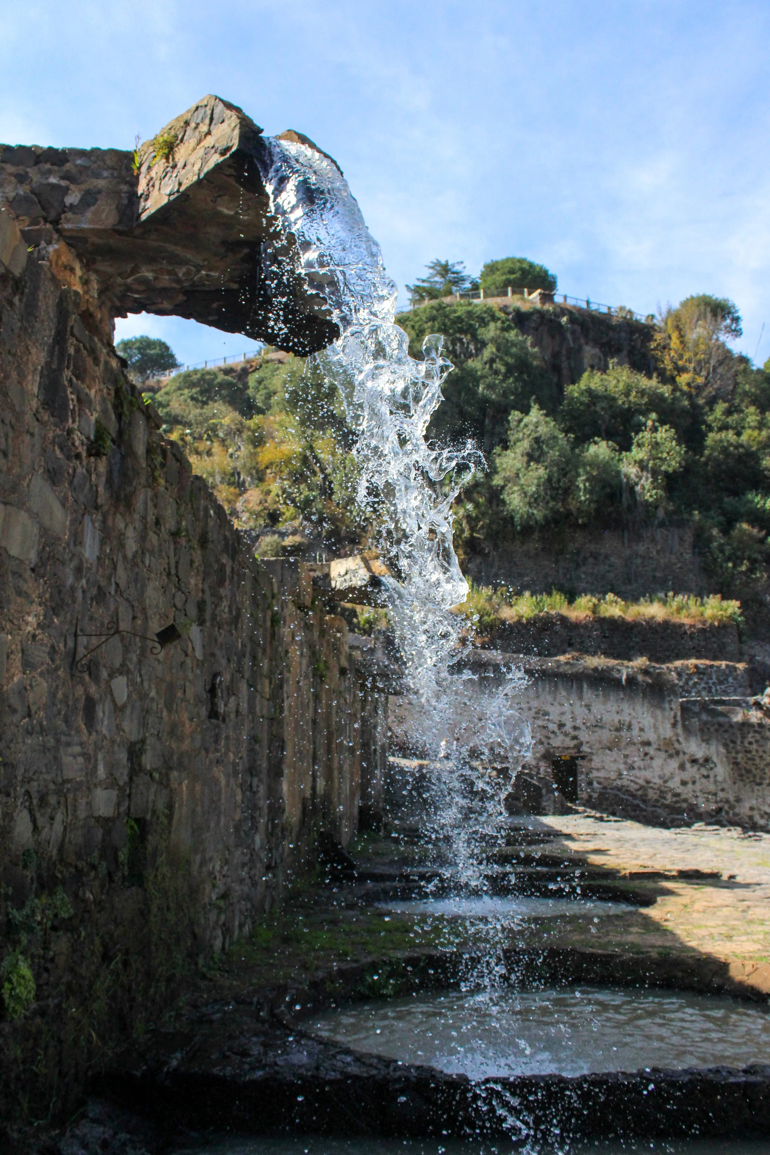 Fuente de agua en una zona con muro de piedra y colinas con vegetación en el fondo