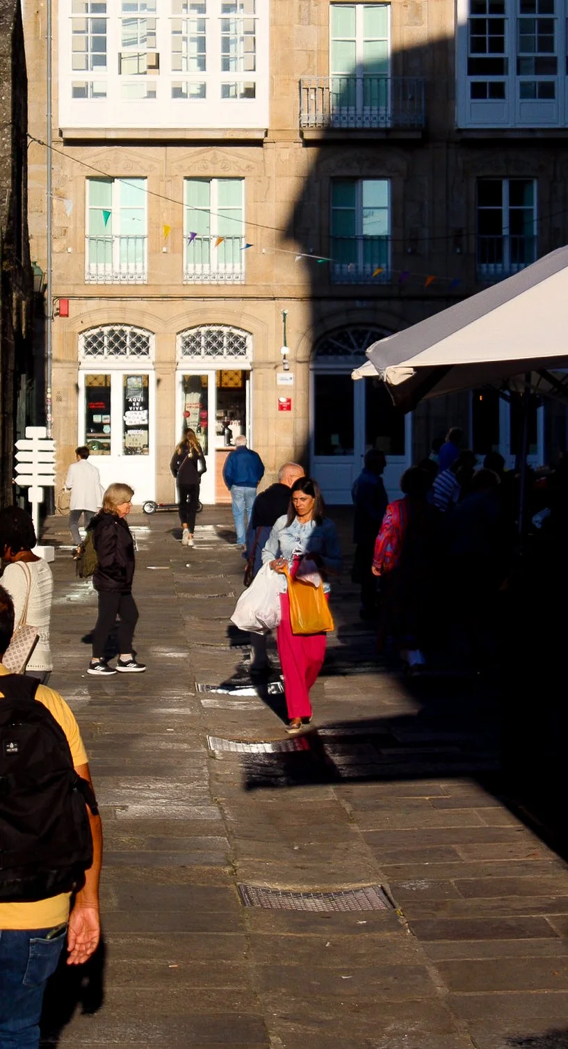 Personas caminando por una calle en una ciudad, con un edificio de fondo y una parte en sombra en la parte superior derecha, además hay una tienda y una terraza con toldo