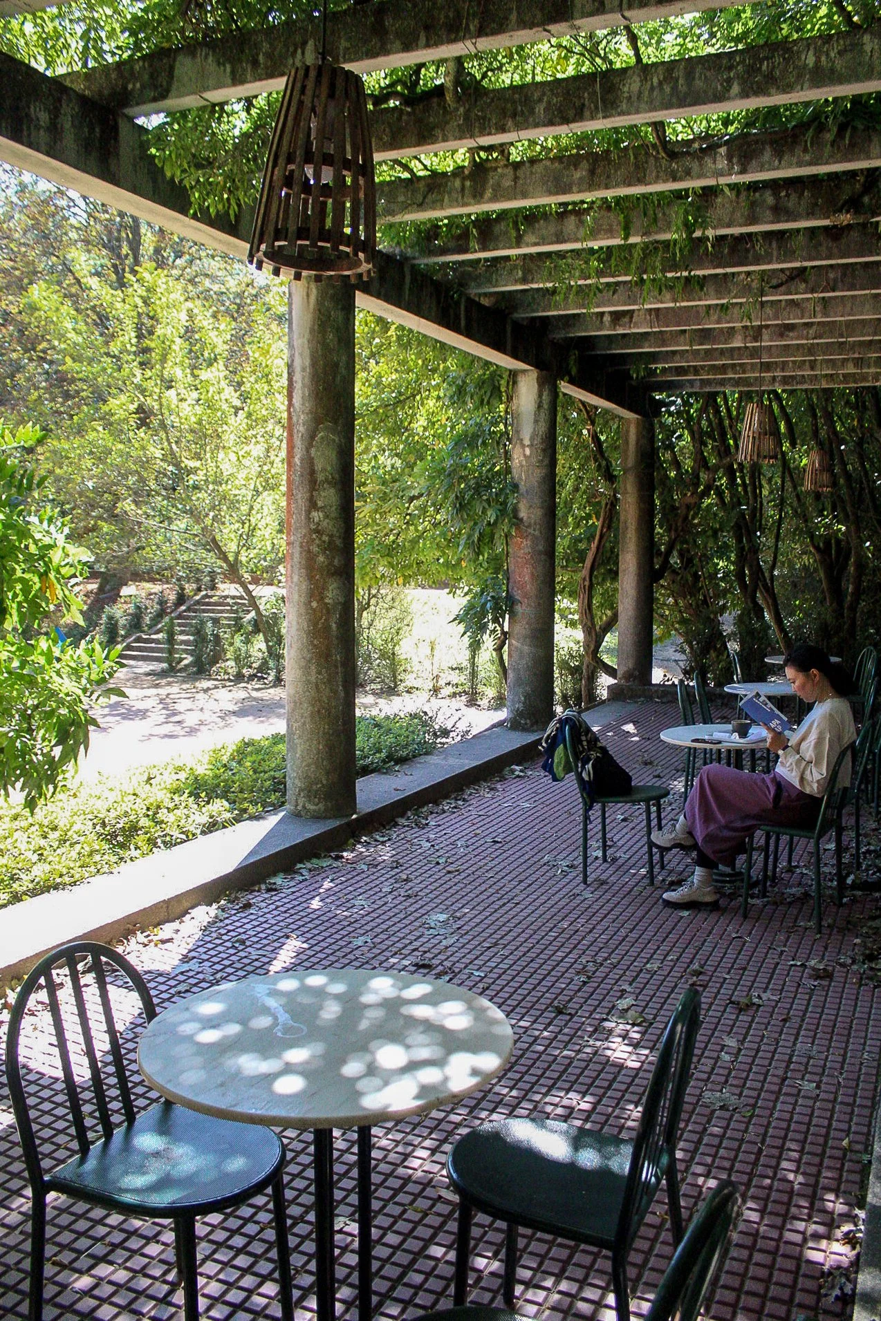Una terraza sombreada con plantas y columnas de cemento, con una mujer sentada leyendo un libro, mesas y sillas de metal, y una lámpara colgante de madera.