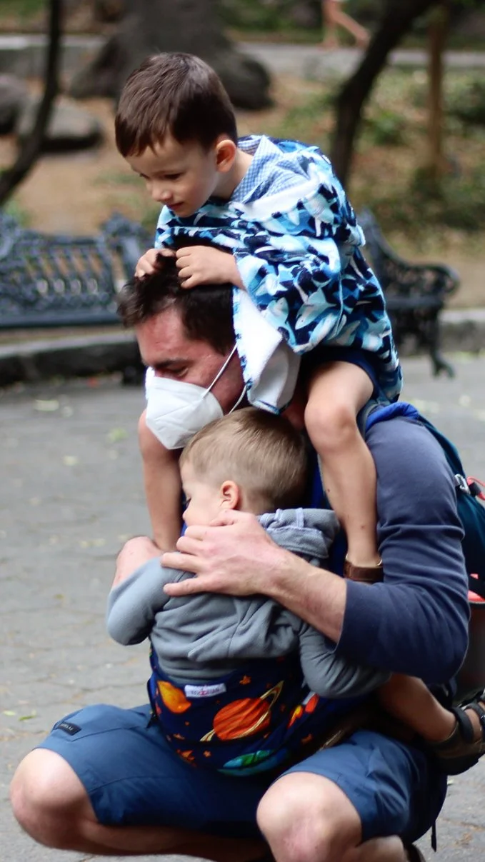 Un hombre con mascarilla jugando con dos niños en un parque, uno sobre sus hombros y otro en sus brazos.