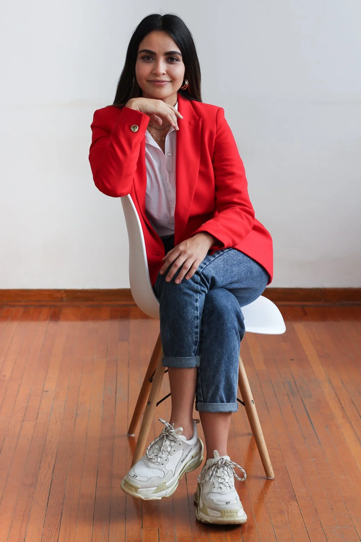 Una mujer sentada en una silla blanca, vestida con un blazer rojo, camiseta blanca, jeans y zapatillas blancas, en un ambiente con piso de madera y pared blanca.