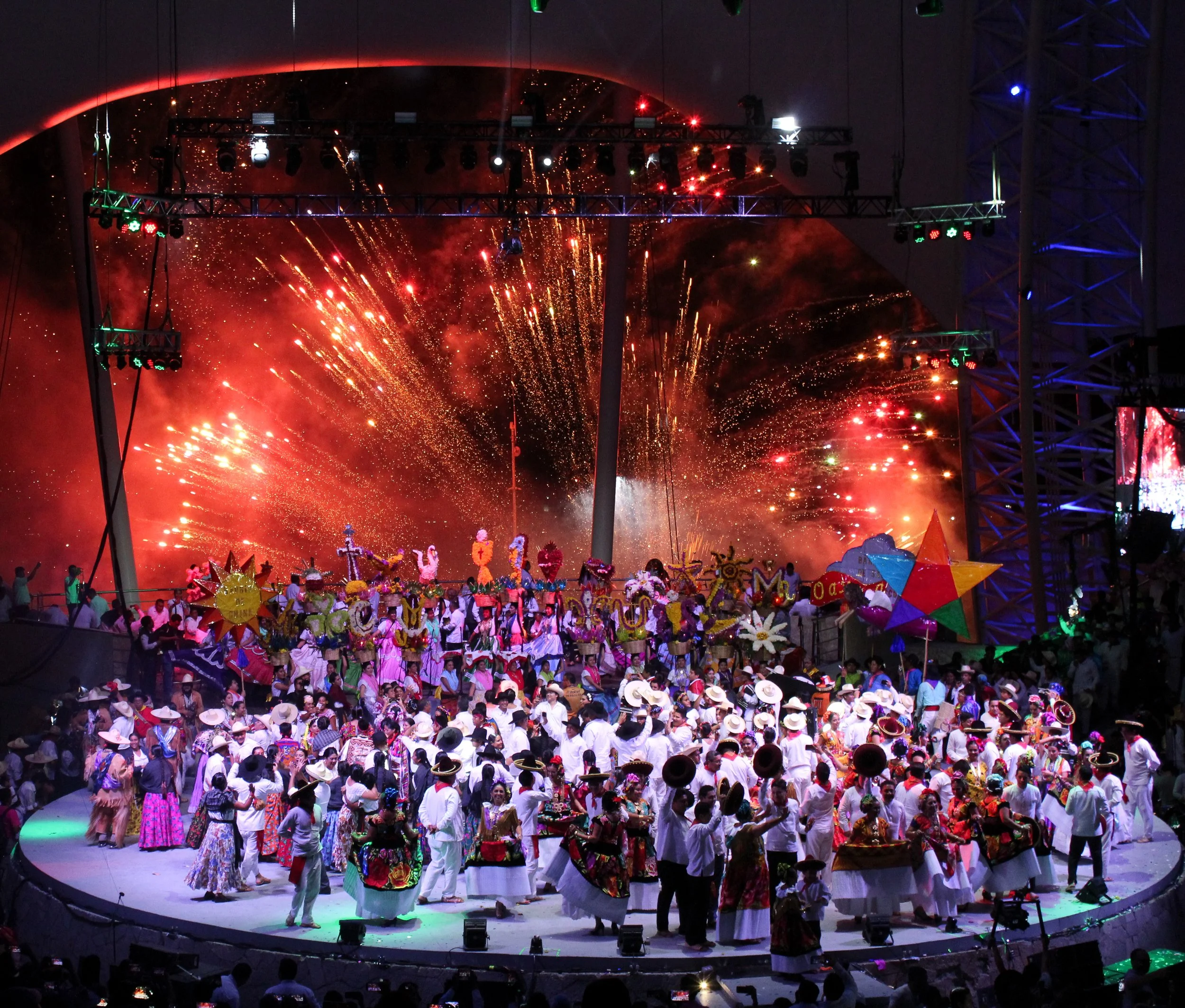 Celebración con fuegos artificiales y un grupo grande de personas vestidas con ropa tradicional y sombreros en un escenario.