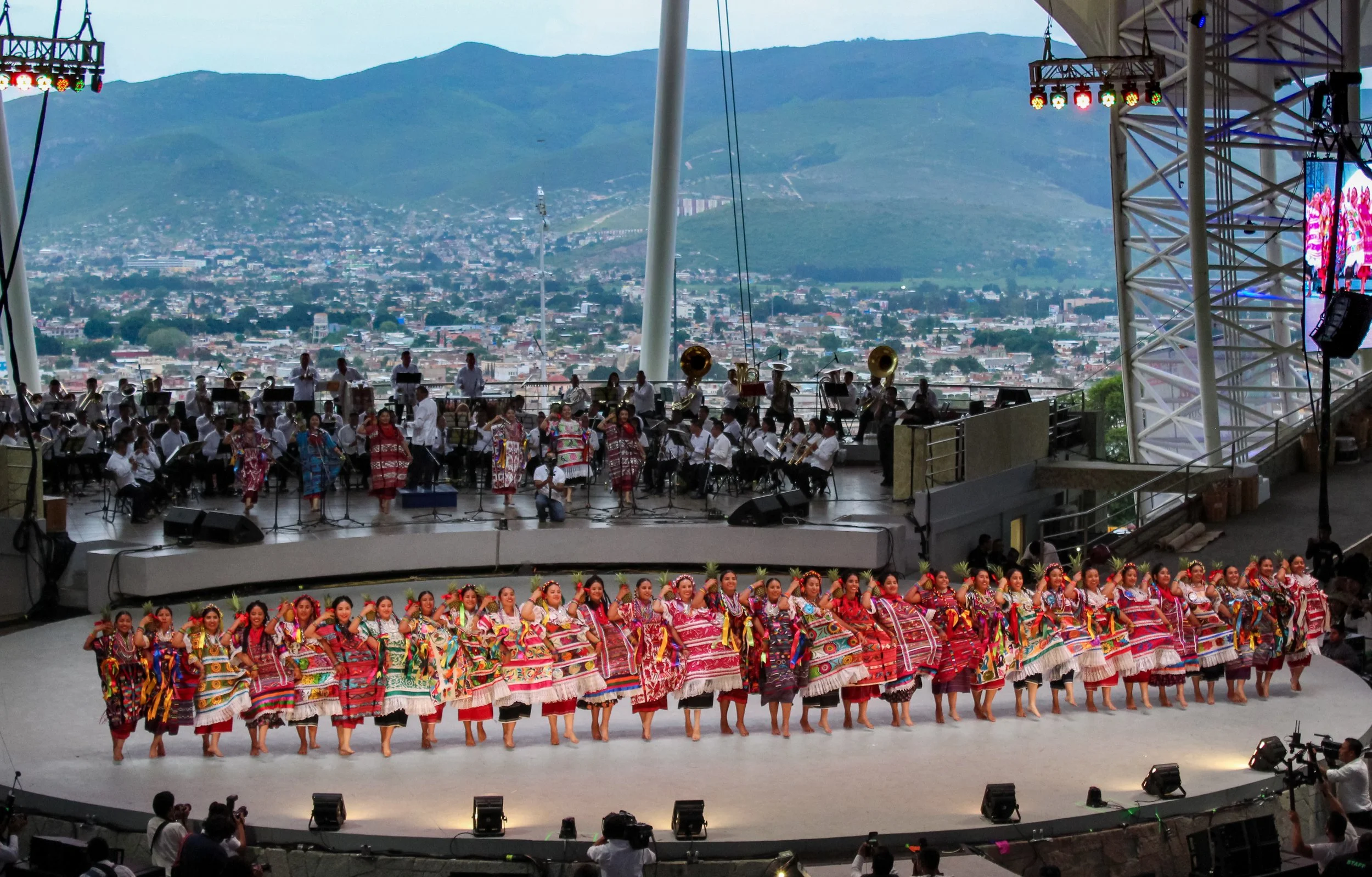 Espectáculo tradicional con baile en un escenario al aire libre, con un grupo de mujeres vestidas con trajes coloridos y diseño folklórico mexicano, al frente y un ensamble musical con instrumentos tradicionales en el fondo, con vista a montañas y ci