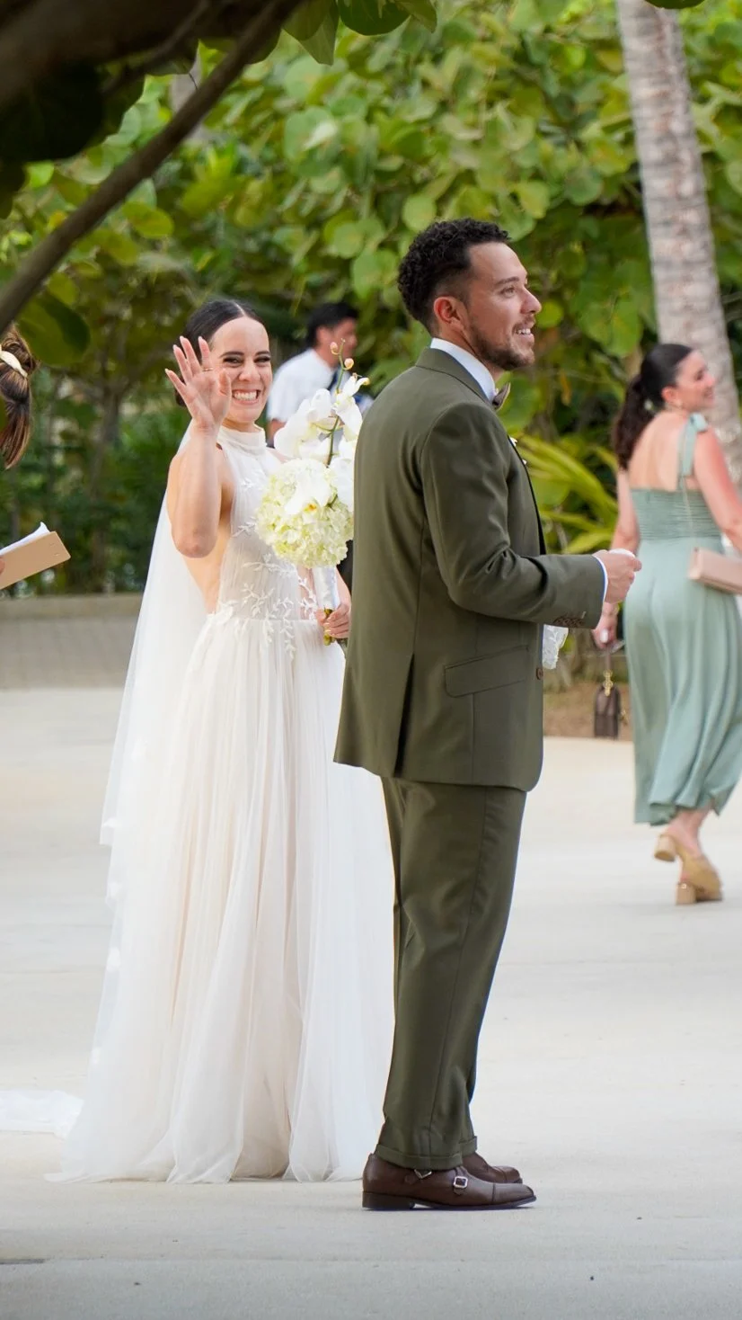 Pareja de recién casados, ella con vestido blanco y ramo, él con saco y corbata, celebrando en un entorno tropical con vegetación y otras personas en el fondo.