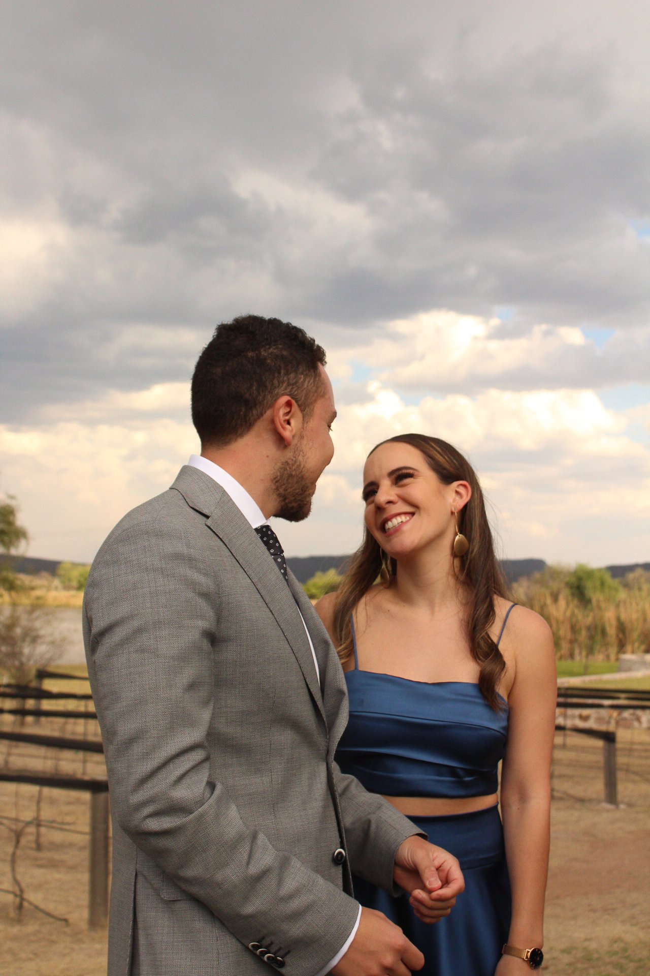 Una pareja sonriendo y mirándose en un entorno al aire libre con cielo nublado y paisaje natural