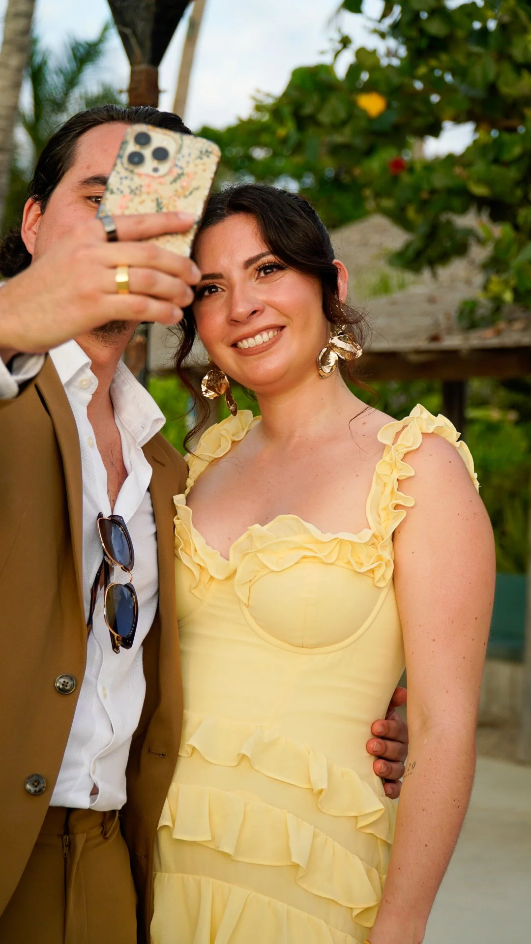 Una mujer y un hombre toman una selfie juntos al aire libre, con fondo de árboles y cielo, la mujer lleva un vestido amarillo con volantes y el hombre viste un saco marrón con gafas colgadas en su camisa.