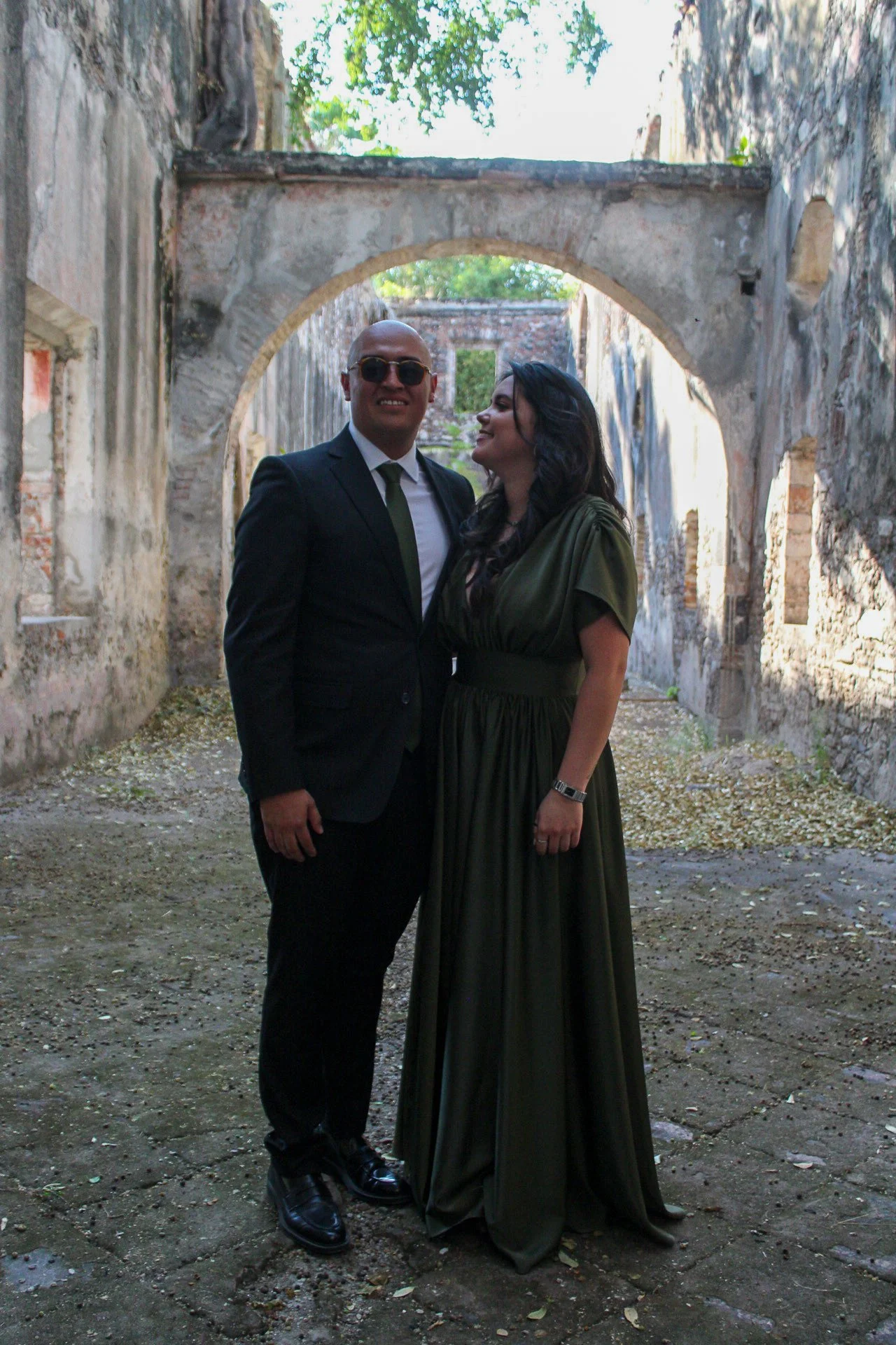 Una pareja vestida elegantemente, el hombre con traje negro y gafas oscuras, la mujer con un vestido largo verde oliva, posando en un lugar con paredes de ladrillo y un arco de piedra en ruinas, con árboles y cielo visible al fondo.