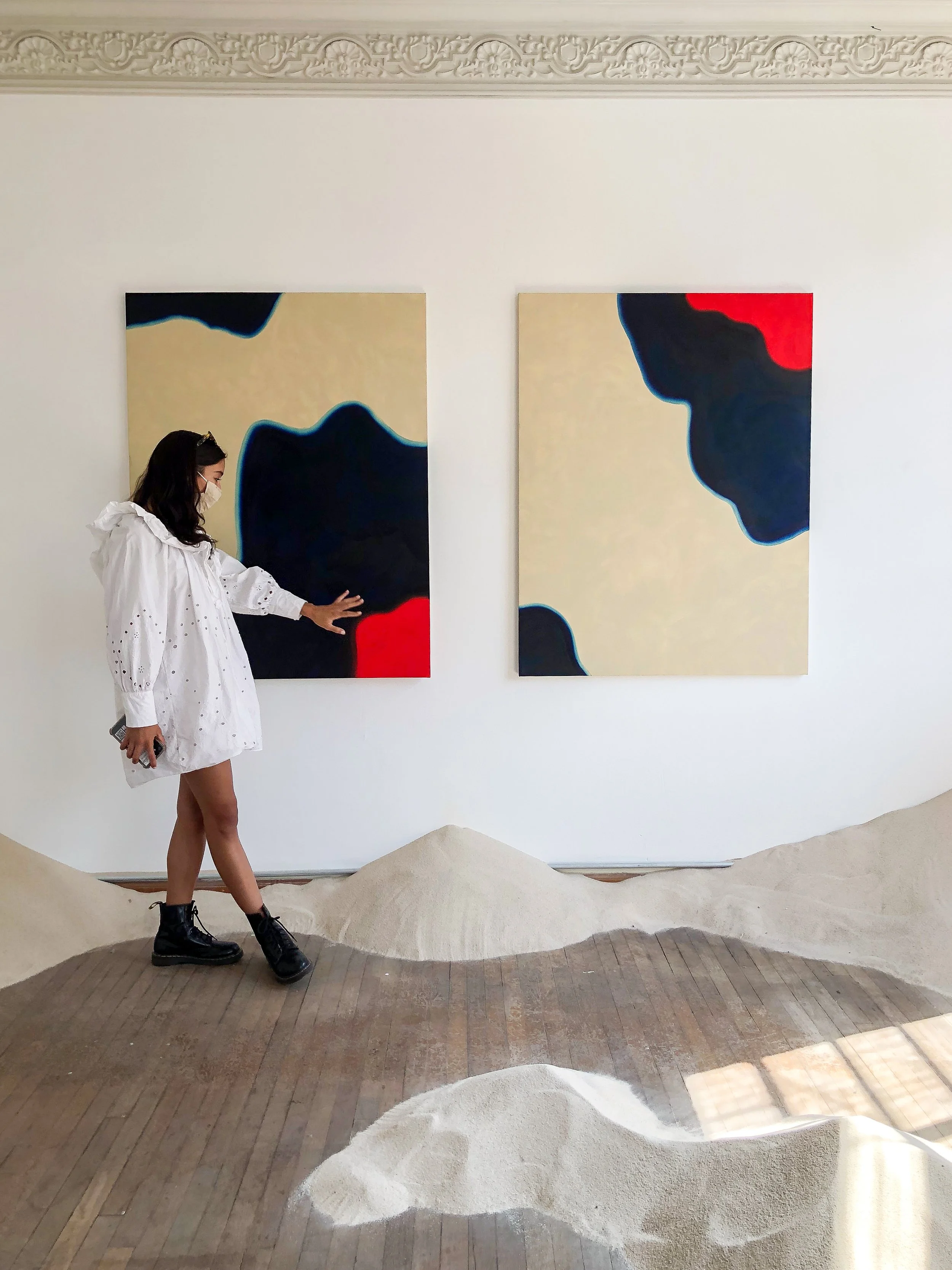 Una mujer con blusa blanca y botas negras en un museo observando dos cuadros abstractos con formas negras, rojas y beige en la pared, con un suelo que simula dunas de arena.