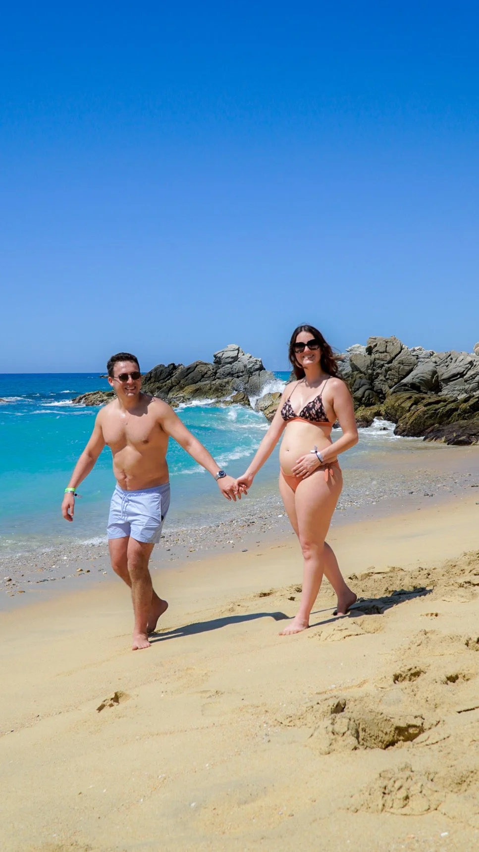 Una pareja caminando por la playa, con rocas y mar al fondo en un día soleado y despejado.