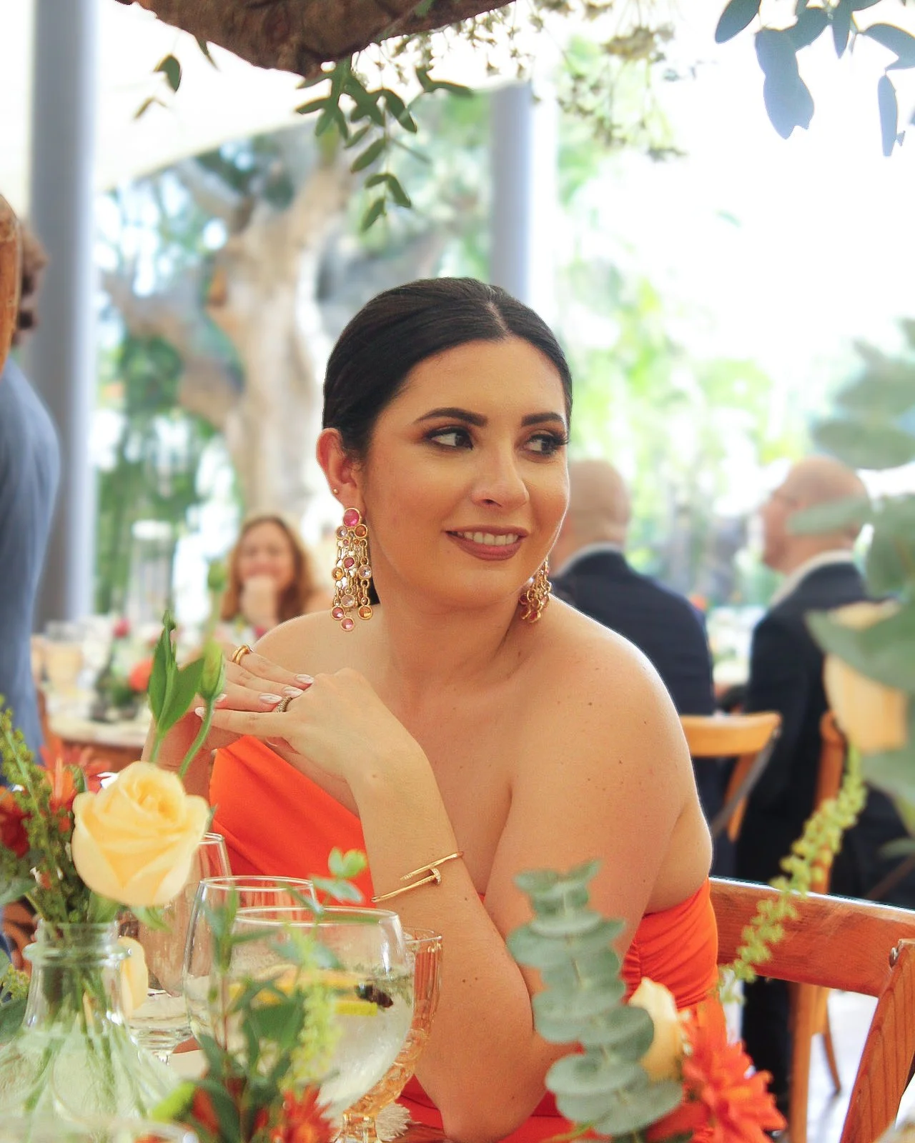 Mujer joven con cabello oscuro, vestido naranja, usando pendientes grandes y dorados, en una celebración en un lugar con mucha luz natural y plantas verdes, rodeada de otras personas en el fondo.