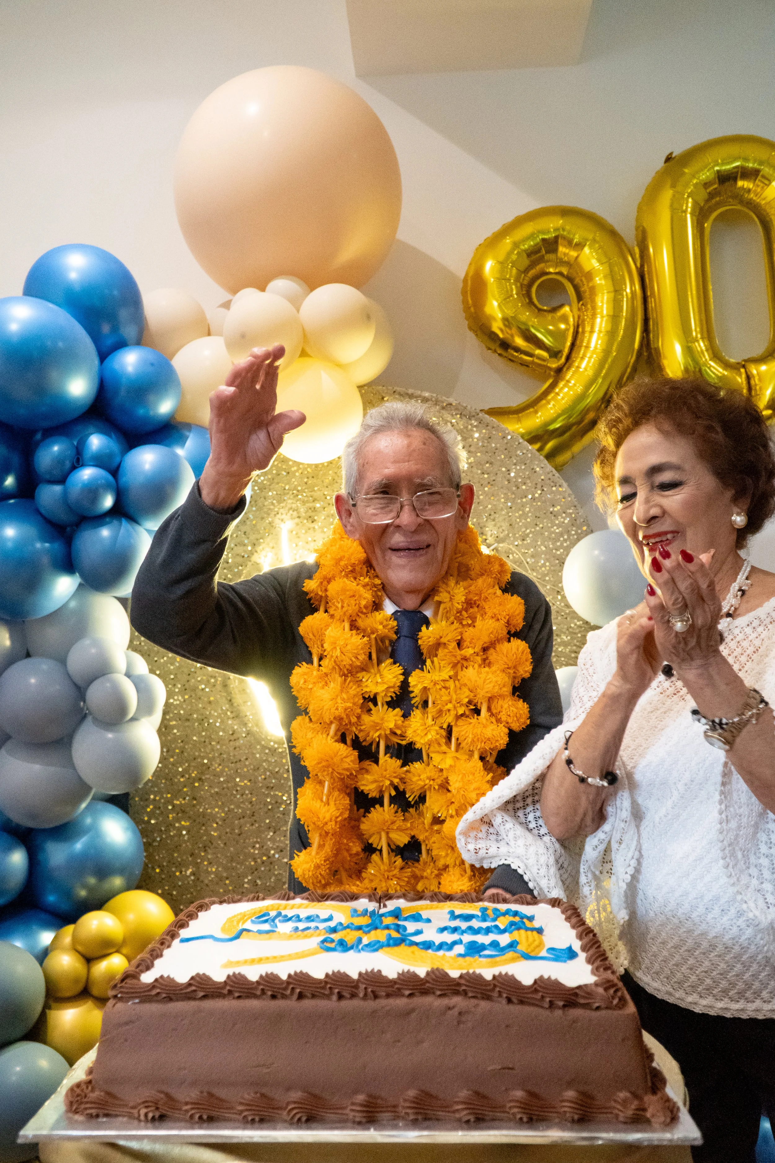 Un hombre y una mujer en una celebración de cumpleaños con decoraciones de globos y un pastel, con globos en forma de números 90.